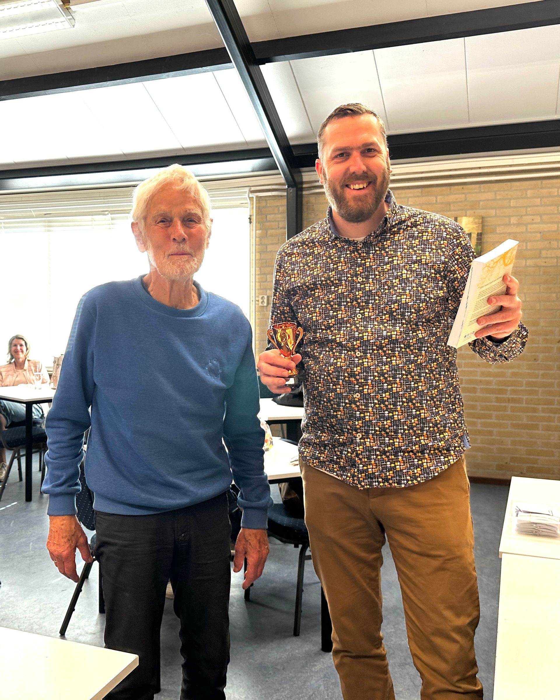 Een man overhandigt een trofee en een boek aan een oudere man; beiden glimlachen binnen, mogelijk tijdens een evenement.