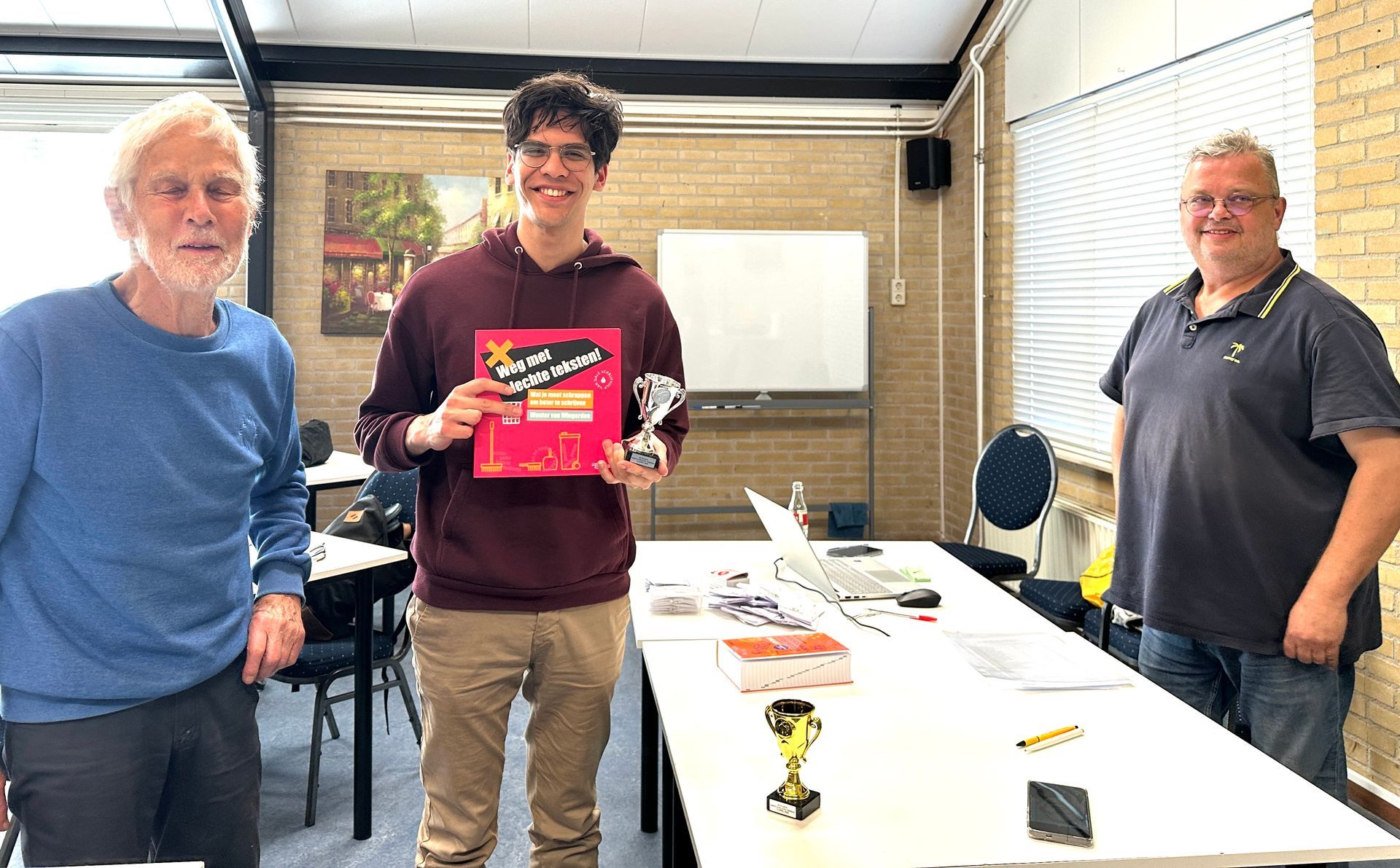 Drie mannen binnen; de middelste houdt een trofee en een boek vast, waarschijnlijk een prijs.