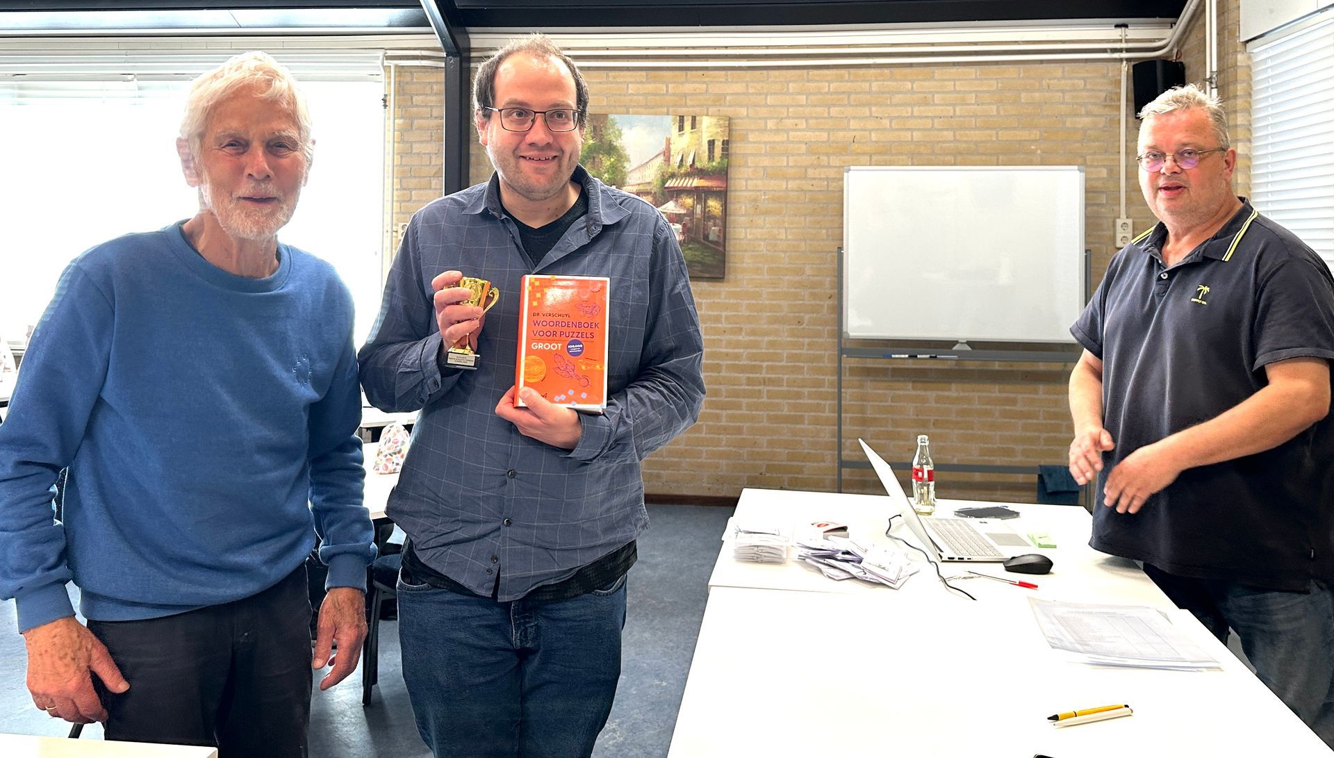 Drie mannen staan, een van hen houdt een boek vast. Ze bevinden zich in een kantooromgeving met een whiteboard en een laptop op een tafel.