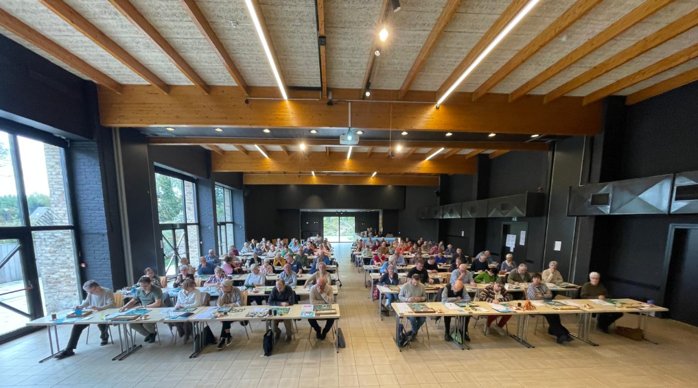 Grote vergaderzaal met veel mensen aan lange tafels; natuurlijk licht, hout en donkere accenten.