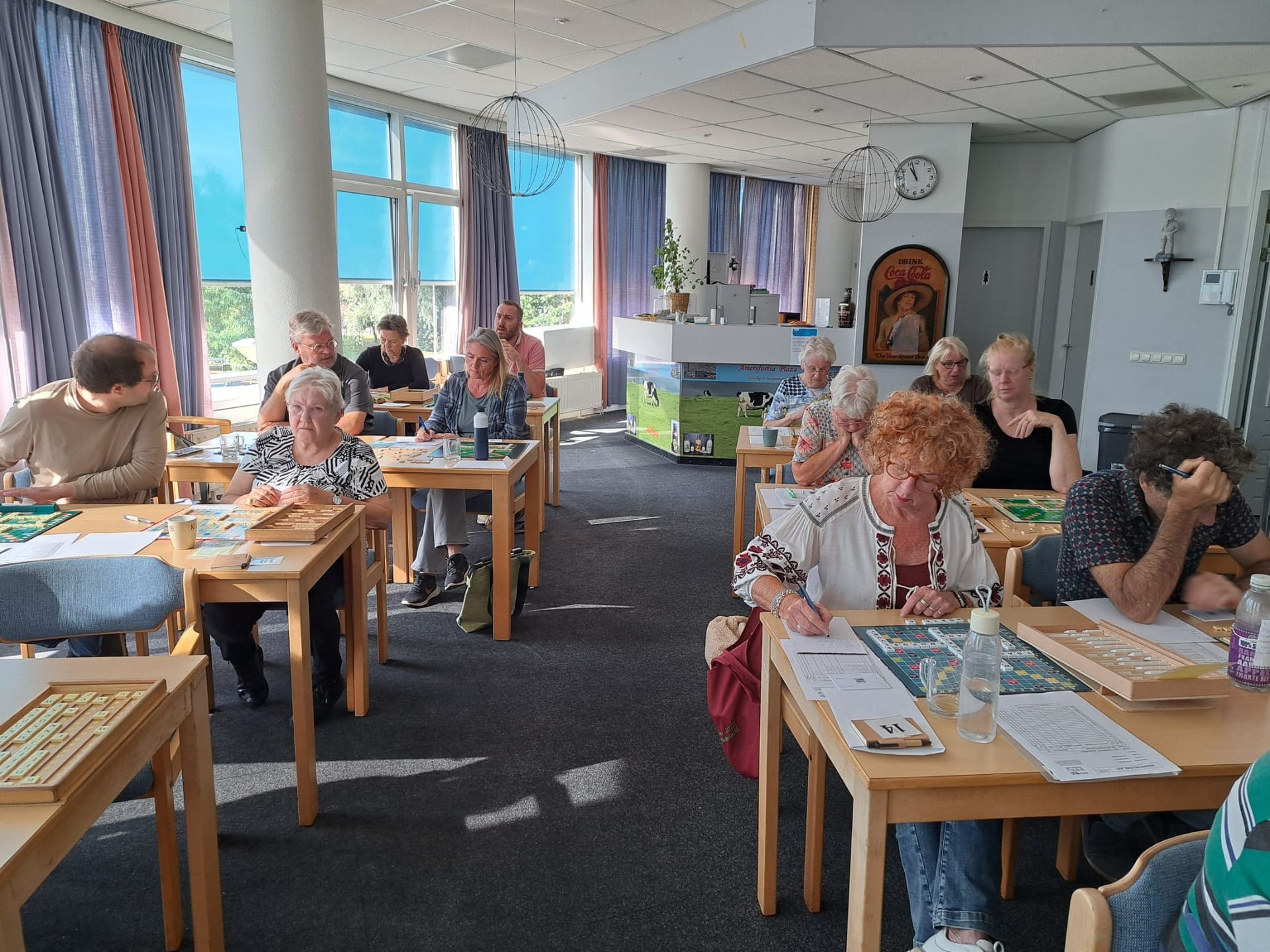 Mensen zitten aan tafels, waarschijnlijk in een klaslokaal, en zijn met een activiteit bezig; ramen zorgen voor natuurlijk licht.