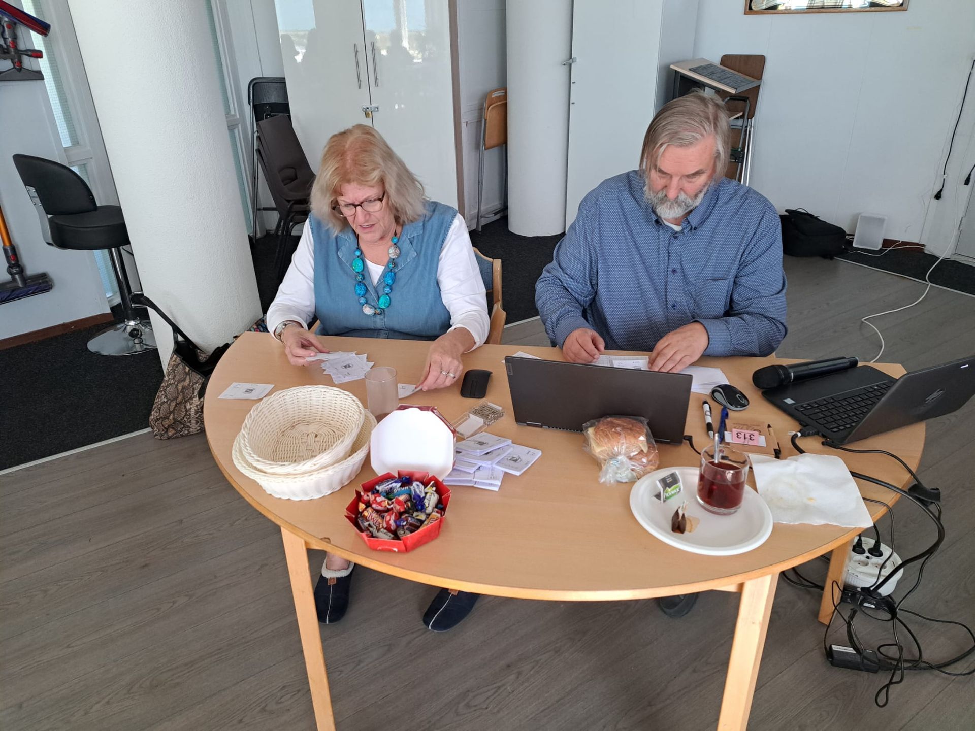 Twee mensen zitten aan een ronde tafel en werken met kaarten en een laptop in een goed verlichte ruimte.