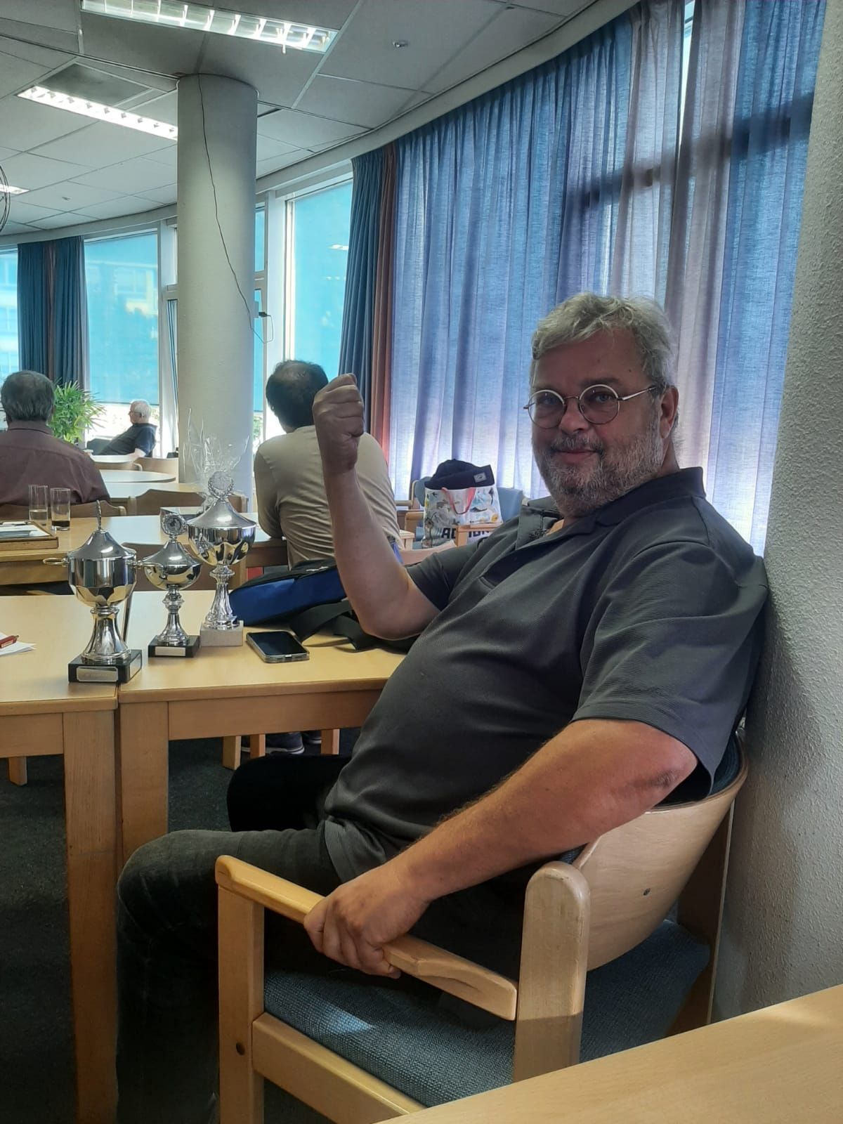 Een man in een grijs shirt steekt zijn duim omhoog en lacht naast trofeeën die binnen op een tafel staan.