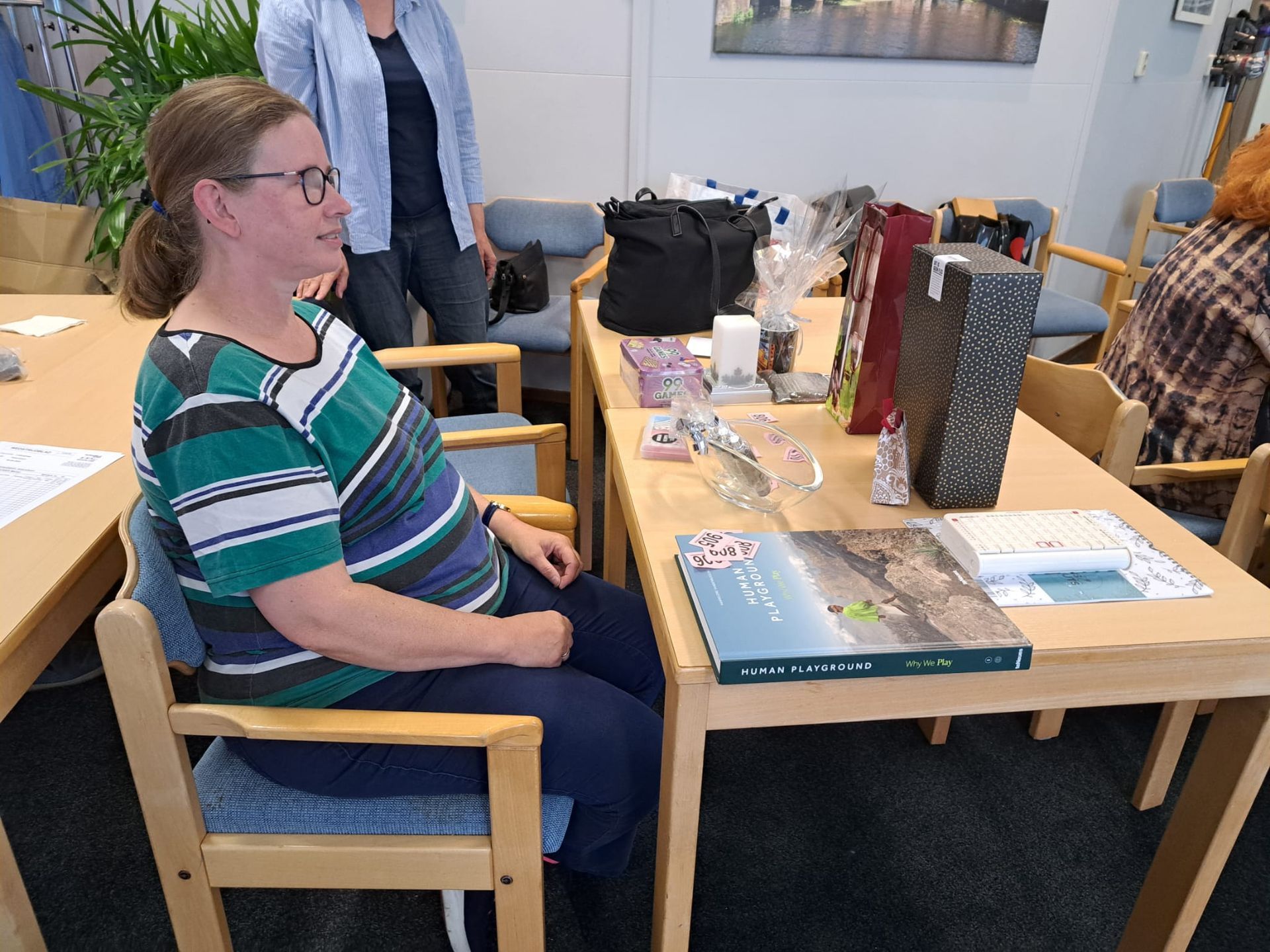 Een vrouw zit aan een tafel met cadeaus, en een andere persoon staat vlakbij in een kamer met ander meubilair.