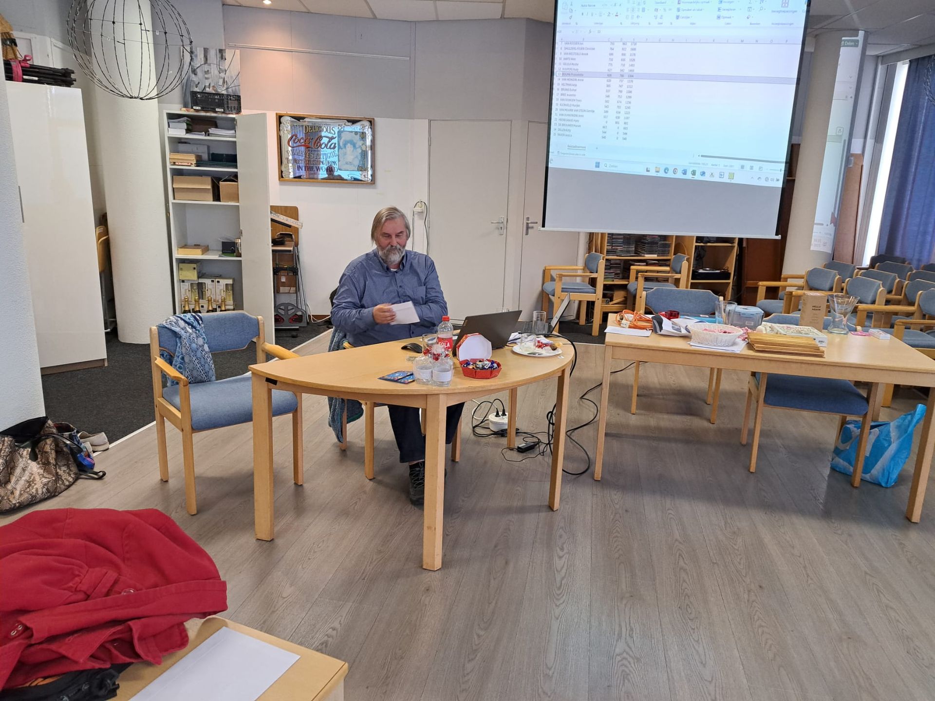 Een man zit aan een tafel met een laptop en geeft een presentatie op een projectiescherm in een kamer met stoelen en versnaperingen.