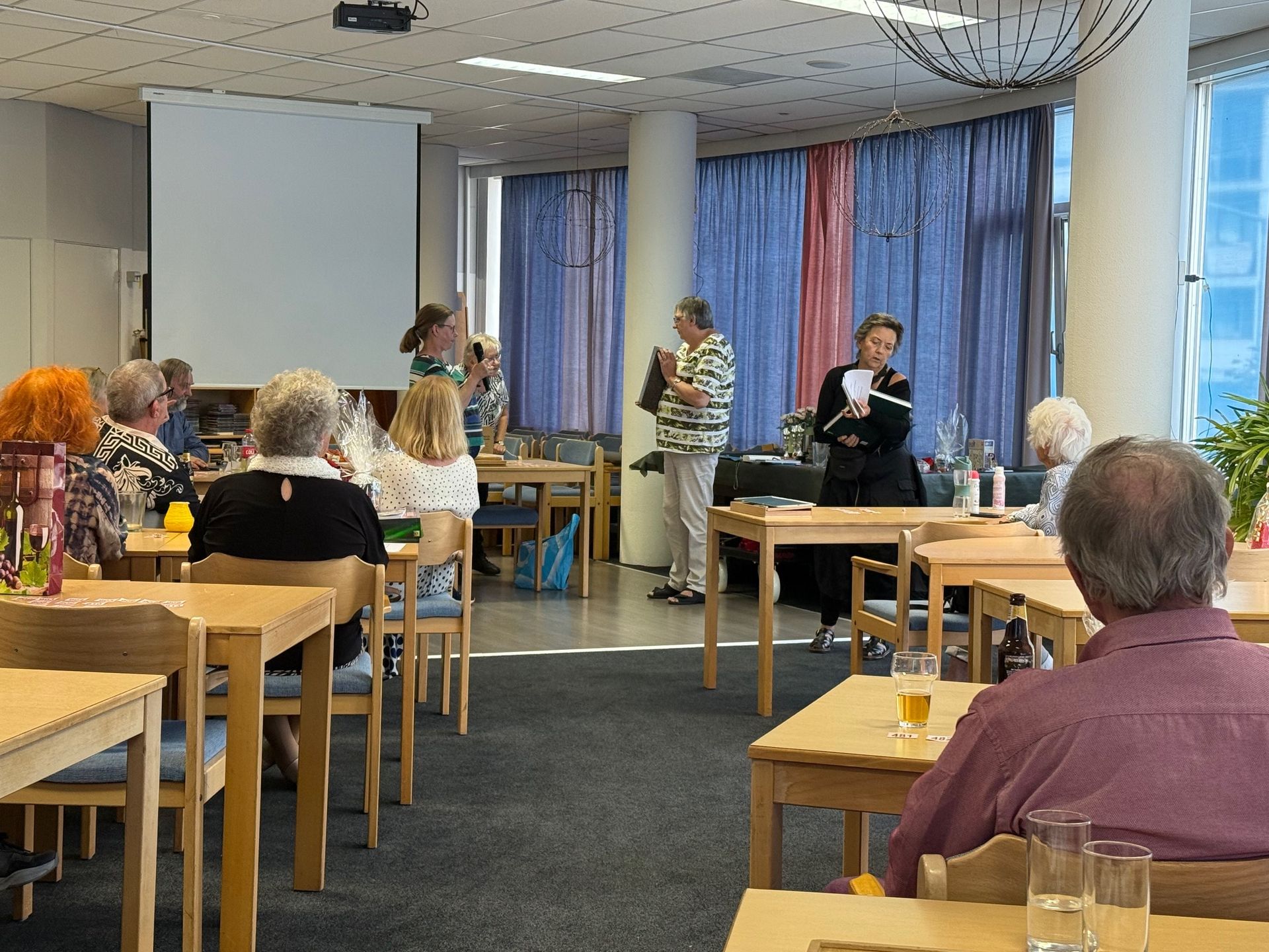 Mensen bevinden zich in een kamer met tafels, sommigen zitten. Een vrouw spreekt aan een tafel met papieren, en een projectiescherm is zichtbaar.