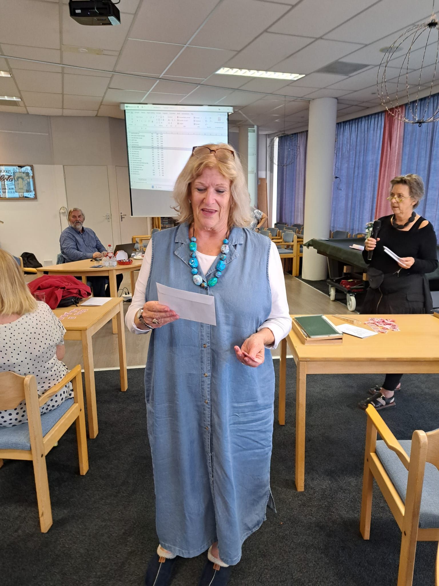 Een vrouw in een spijkerjurk spreekt op een evenement en houdt een document vast. Een andere vrouw staat met een microfoon. Tafels en stoelen.