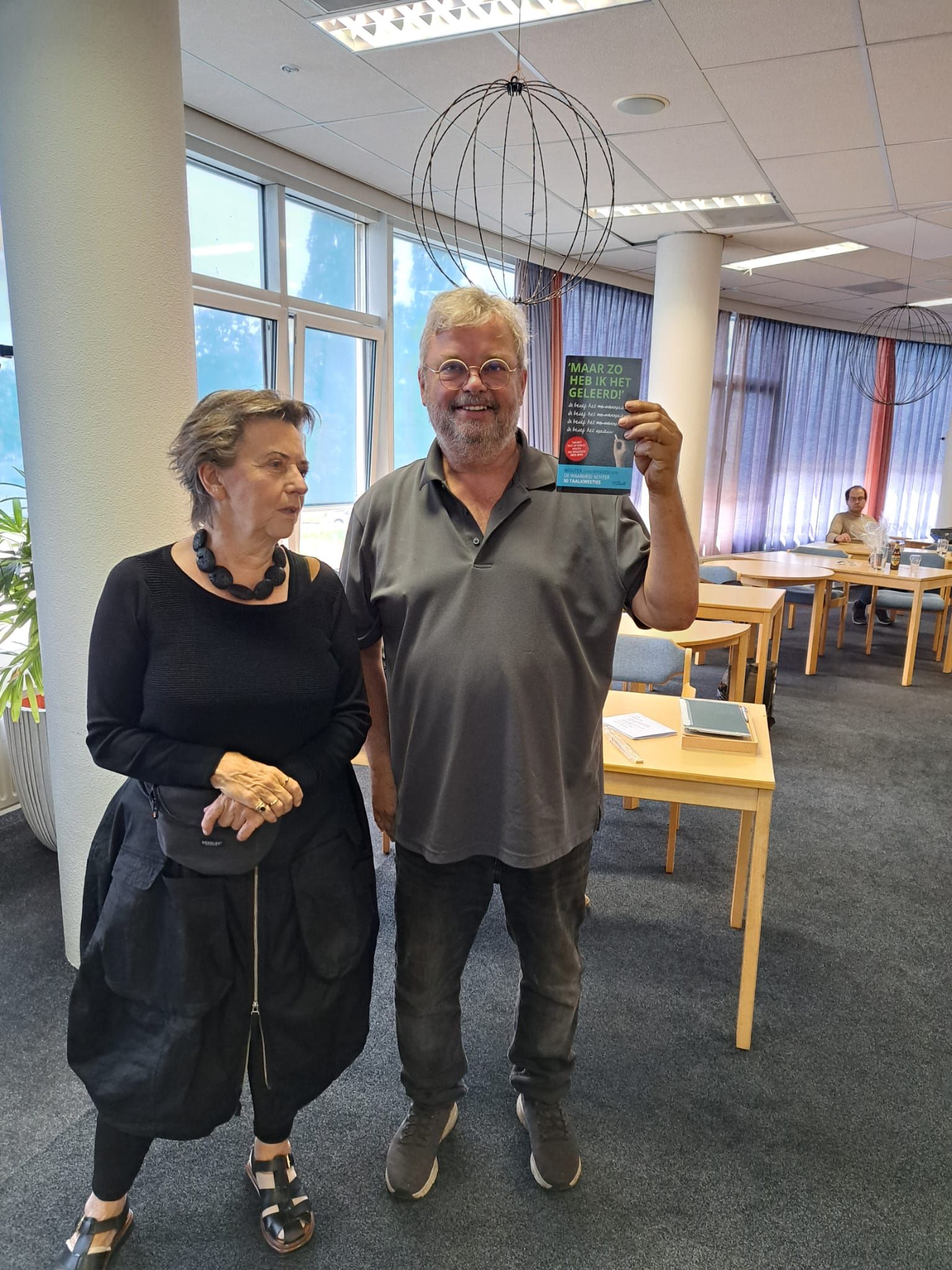 Een man met een boek in zijn hand glimlacht naast een vrouw binnenshuis, bij sfeervol licht.