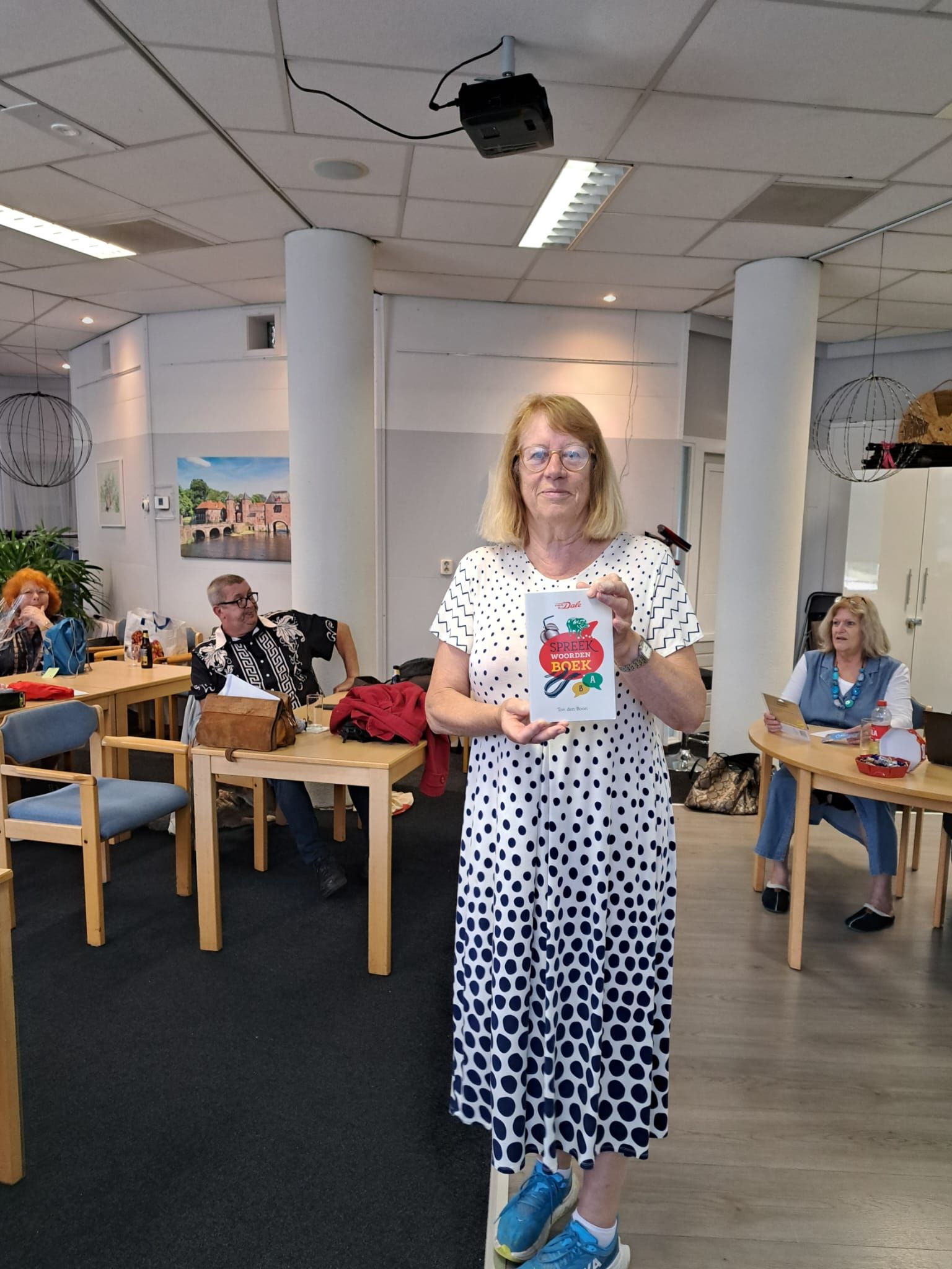 Een vrouw met een kleurrijk boek in haar handen staat in een kamer waar andere mensen aan tafels zitten.