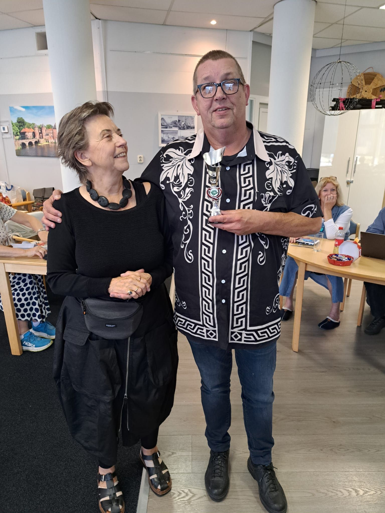 Een vrouw in zwart en een man in een overhemd met patroon houden een prijs vast en glimlachen in een gebouw.