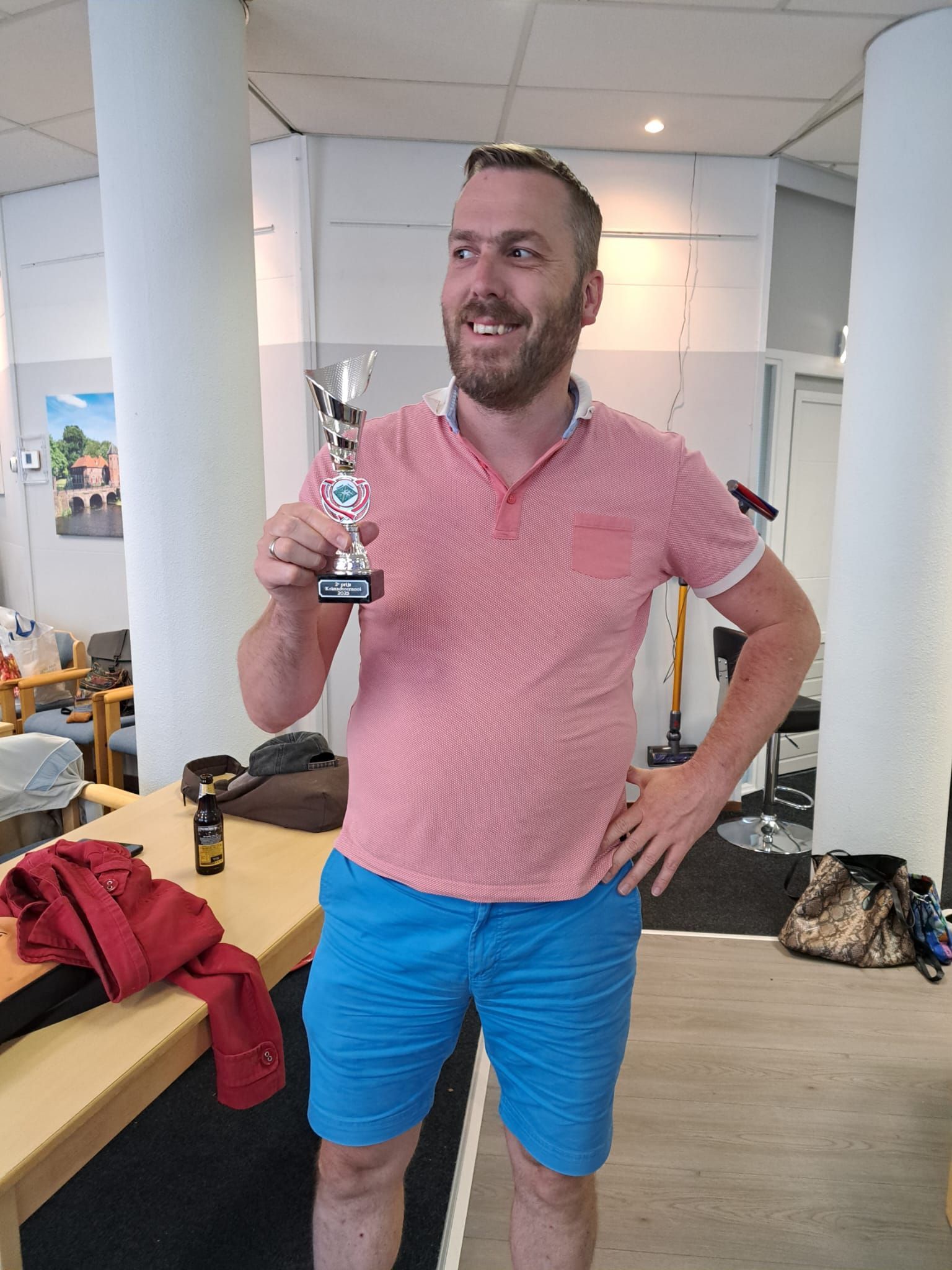 Een man met een trofee in zijn hand, gekleed in een roze shirt en een blauwe korte broek, lacht. Binnen, vlakbij een tafel.