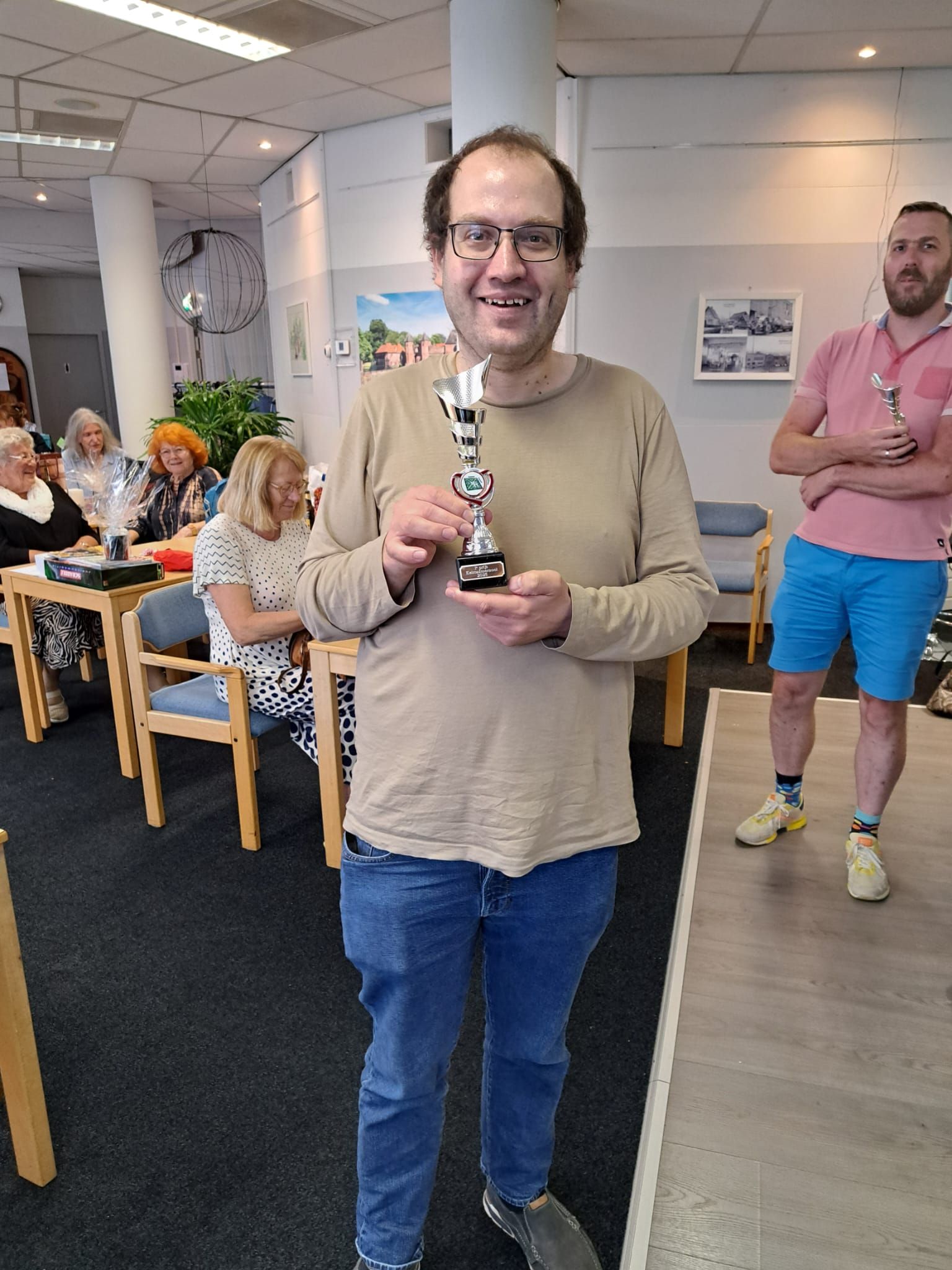 Een man met een trofee in zijn hand lacht. Hij draagt ​​een spijkerbroek en een beige shirt en staat binnen tussen andere mensen en tafels.