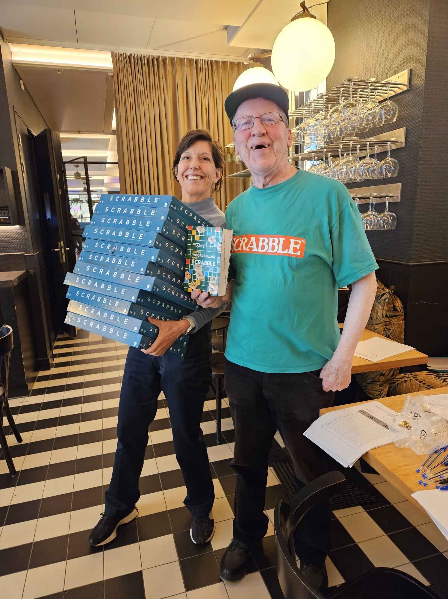 Een vrouw en een man, die stapels Scrabble-speldozen vasthouden, glimlachen in een restaurant.
