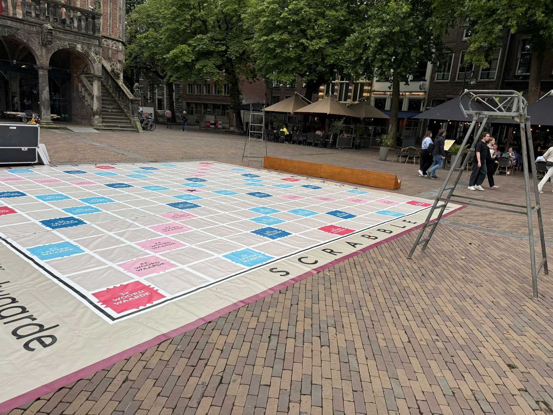 Groot bordspel in de openlucht op een openbaar plein. Gebouwen en mensen op de achtergrond. Roze, blauwe en witte vakjes.