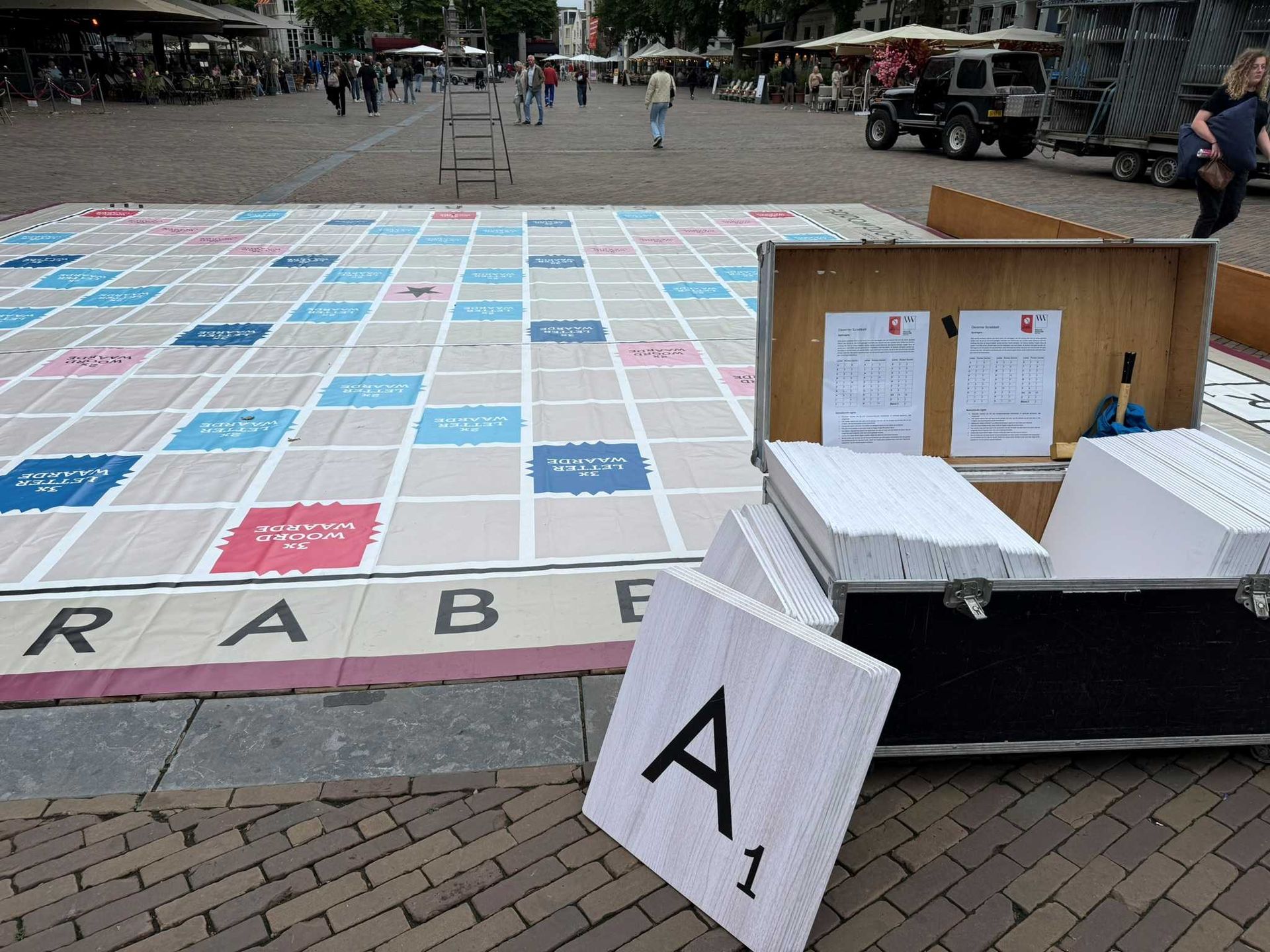 Een gigantisch Scrabble-spel is buiten opgesteld op een stenen plein, met een groot speelbord en een doos met tegels.