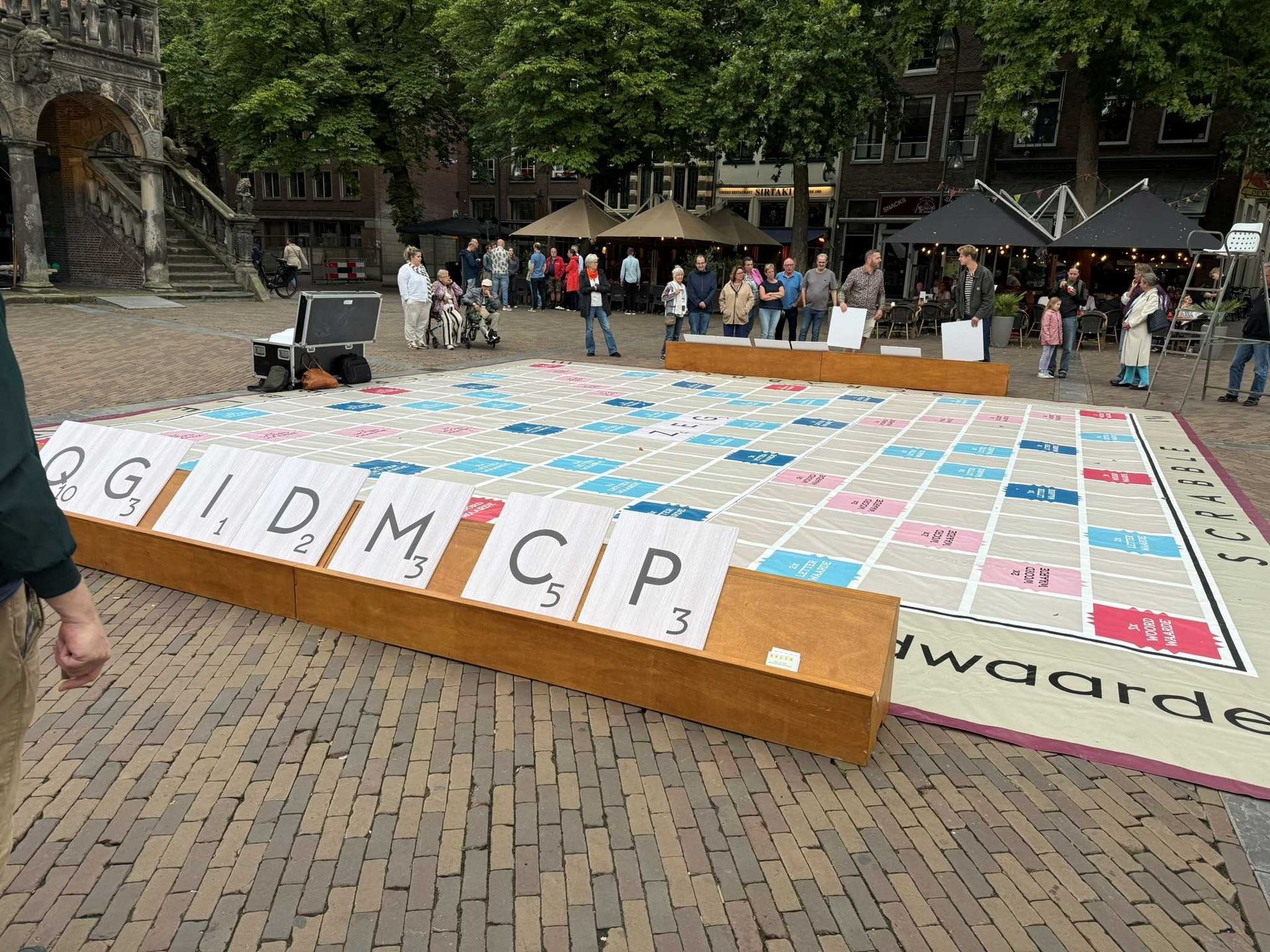 Een groot Scrabble-spel in de openlucht op een stenen plein, met spelers en toeschouwers in de buurt.