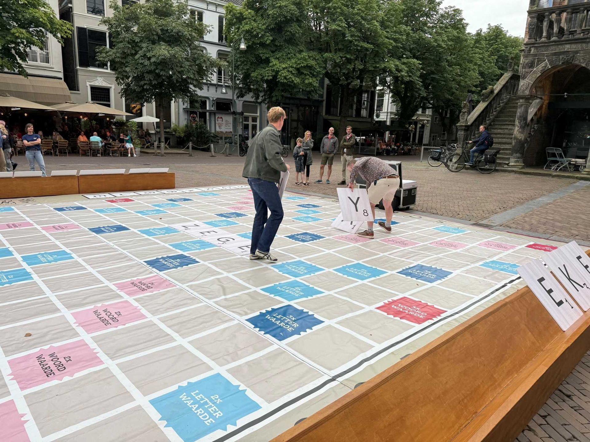 Twee mannen spelen een gigantisch Scrabble-spel op een stadsplein, terwijl andere mensen toekijken.