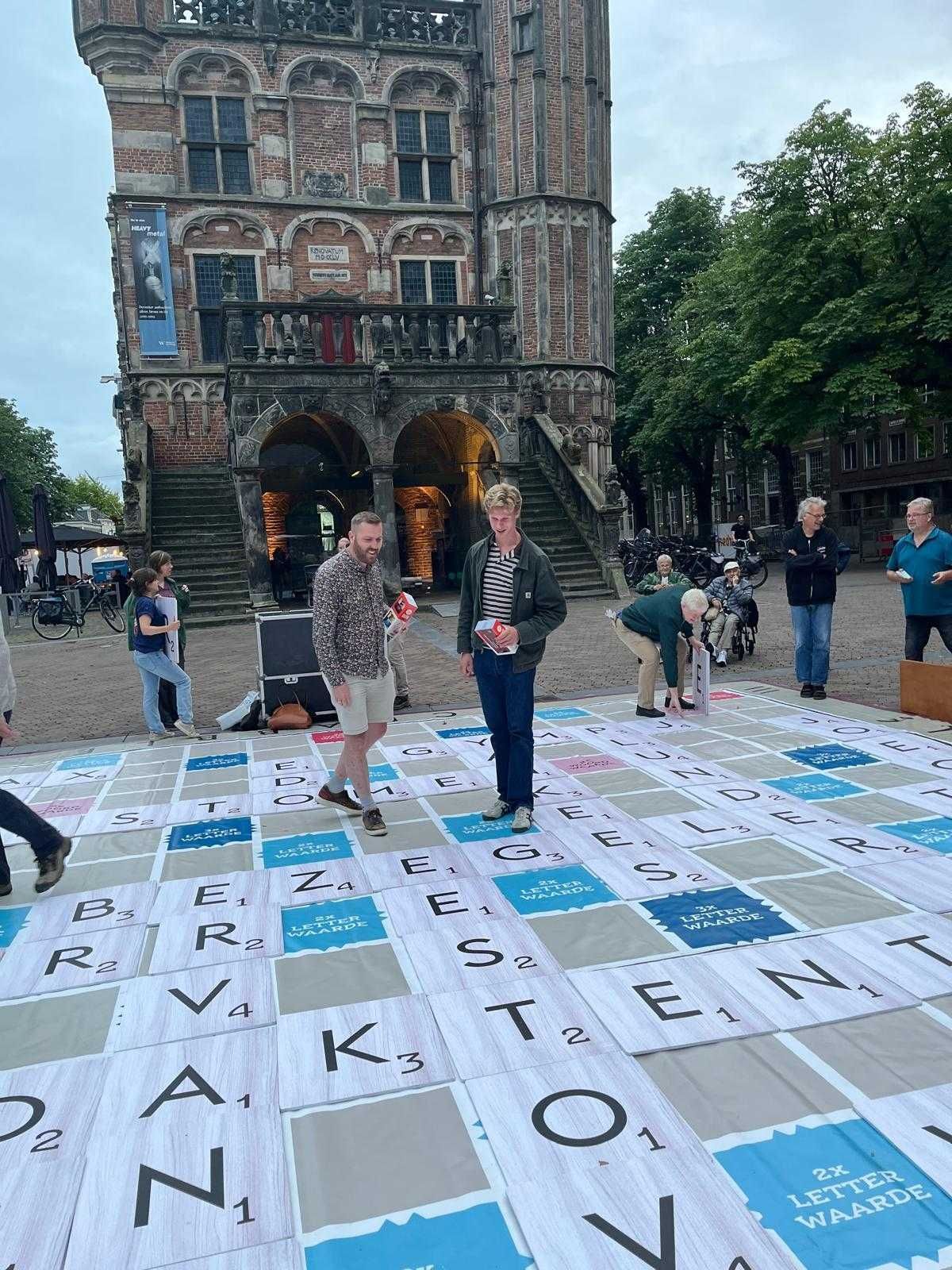 Mensen spelen een gigantisch woordspel op een vierkant tegelbord voor een historisch gebouw.