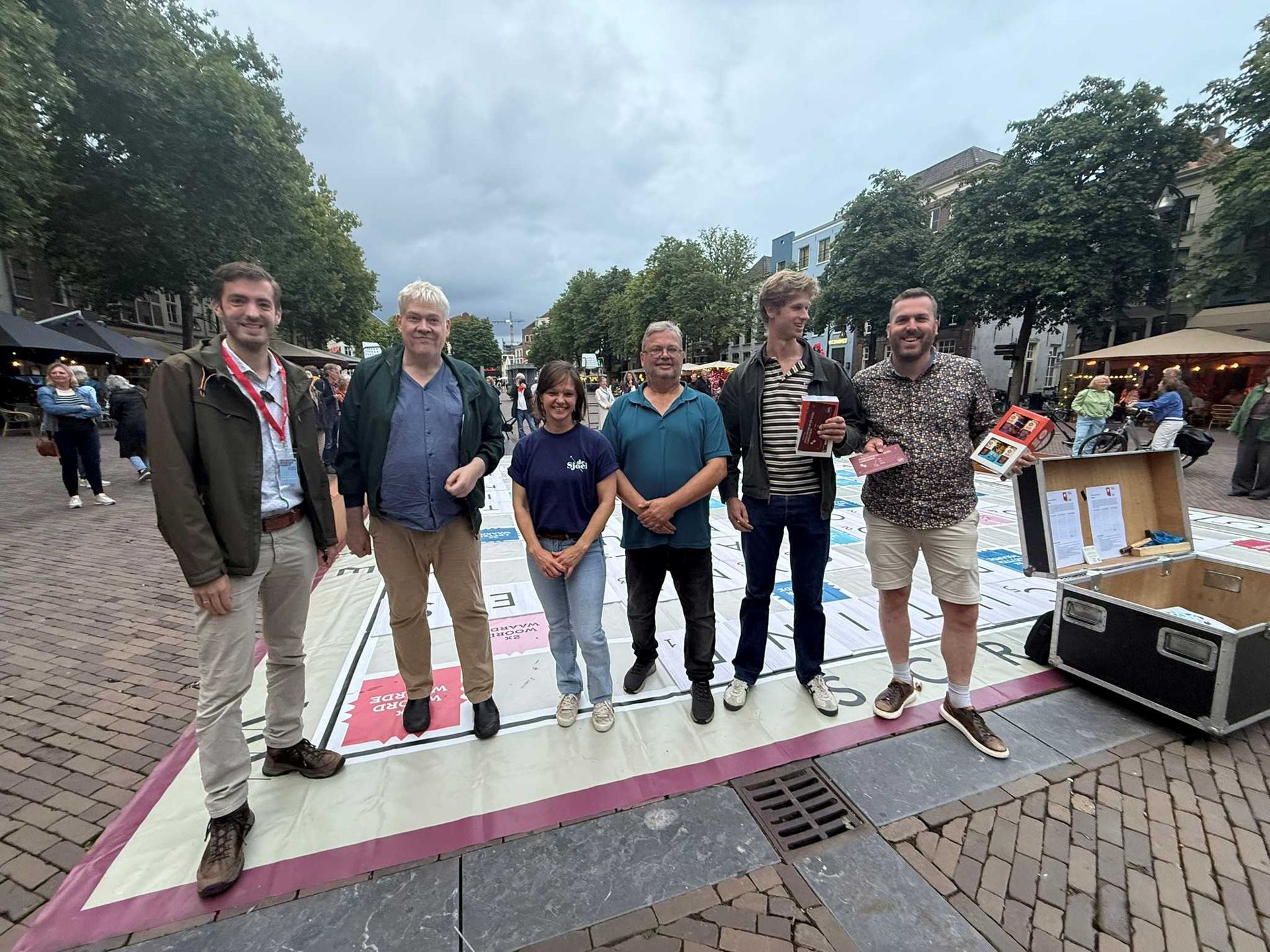 Een groep van zes mensen staat op een gigantisch Monopoly-bord op een stadsplein.