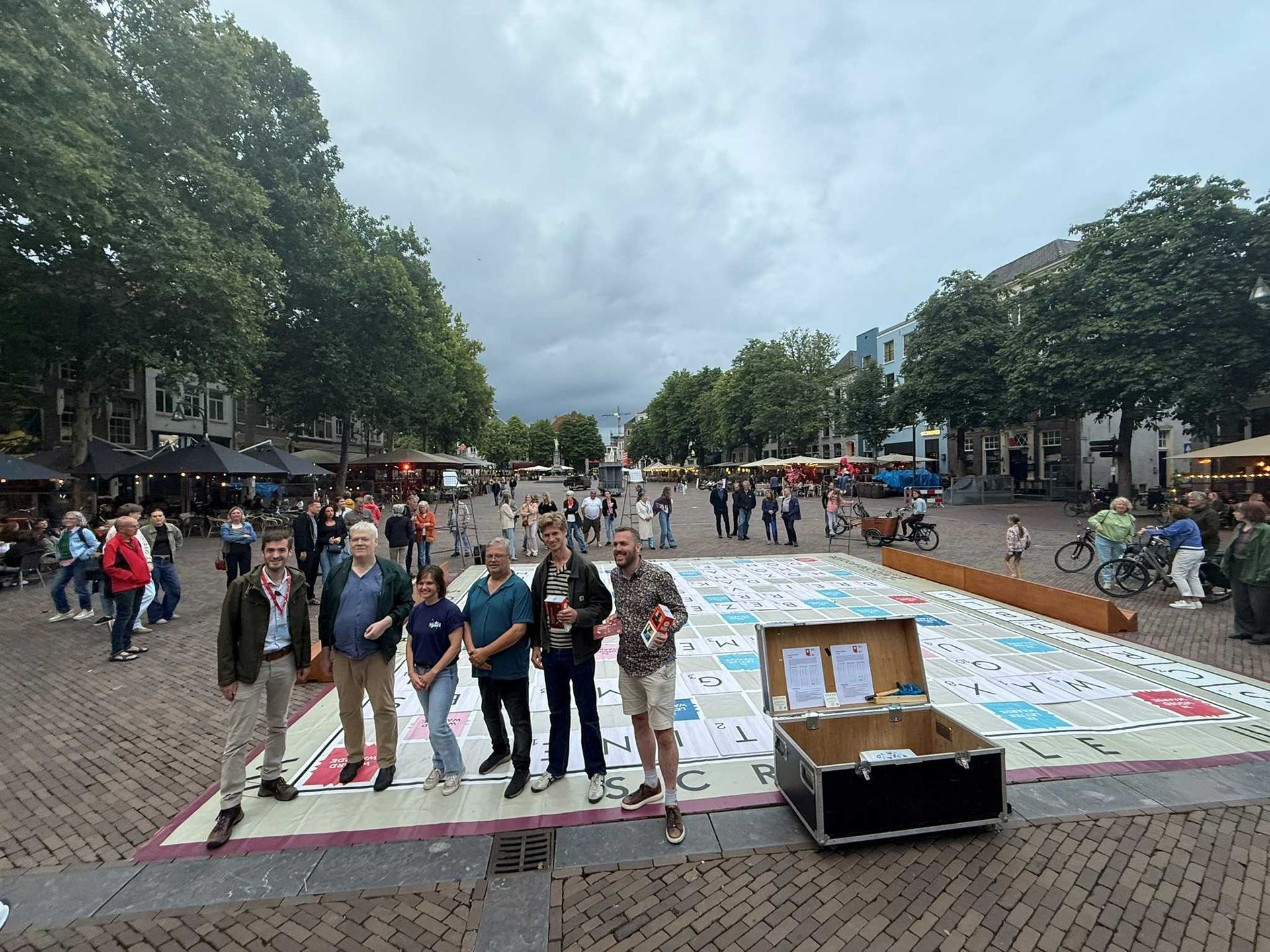 Mensen staan ​​bij een groot spelbord op een openbaar plein. Ernaast staat een doos. Bewolkte lucht.