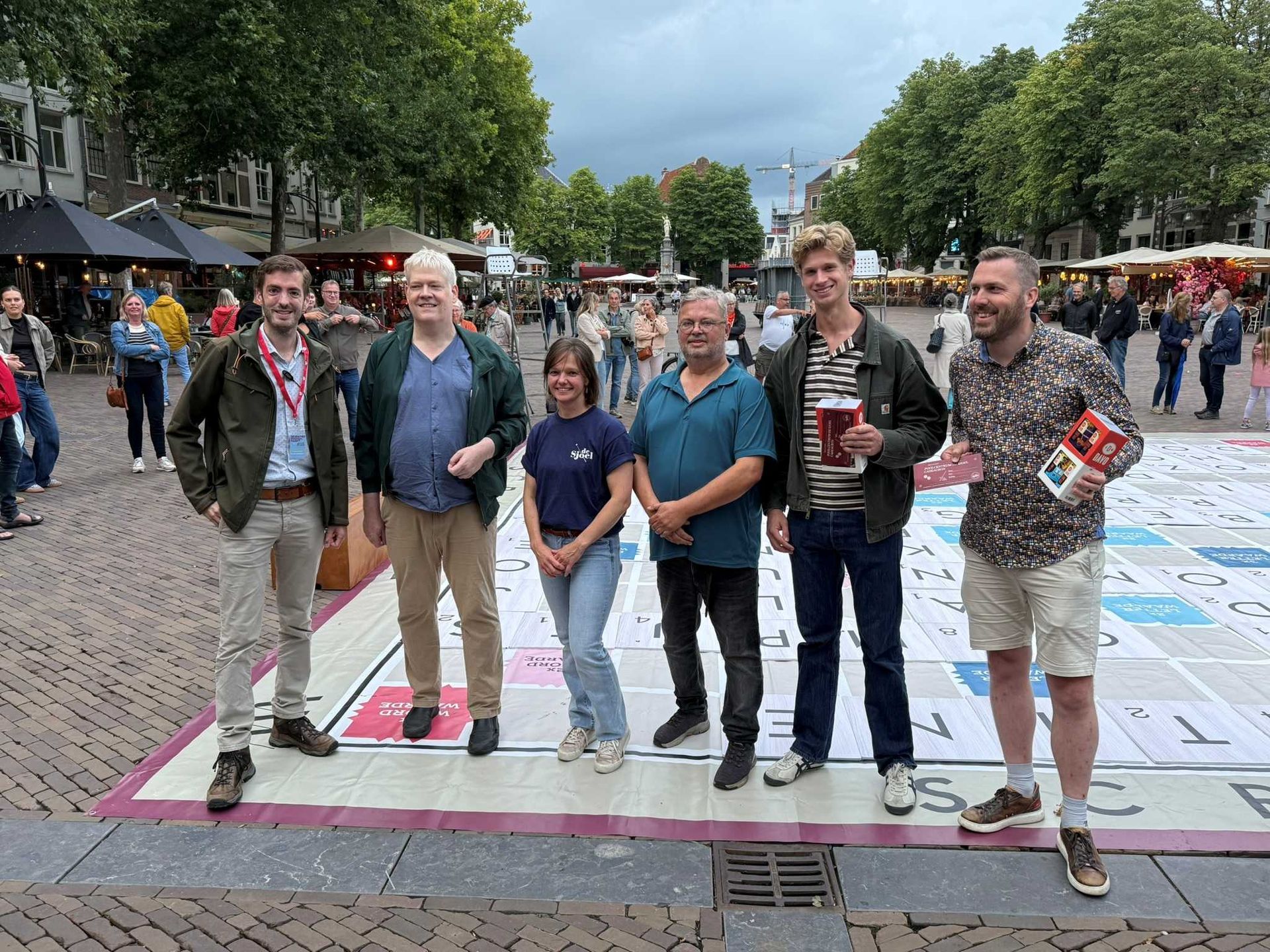 Zes mensen staan ​​op een groot spelbord op een openbaar plein. Ze glimlachen, houden boeken vast en kijken naar de camera.