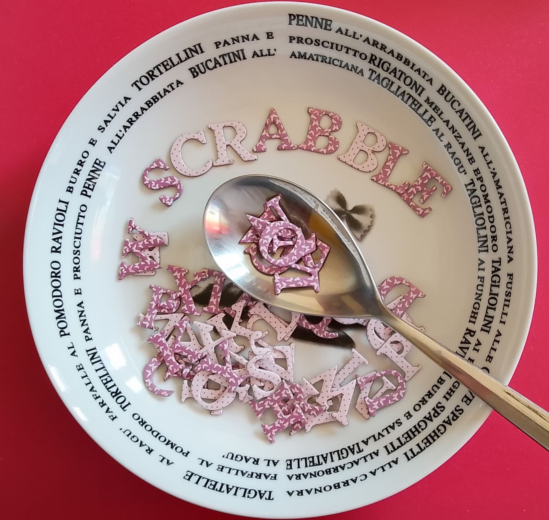 Roze glitter Scrabble-letters in een kom met een lepel; rode achtergrond.