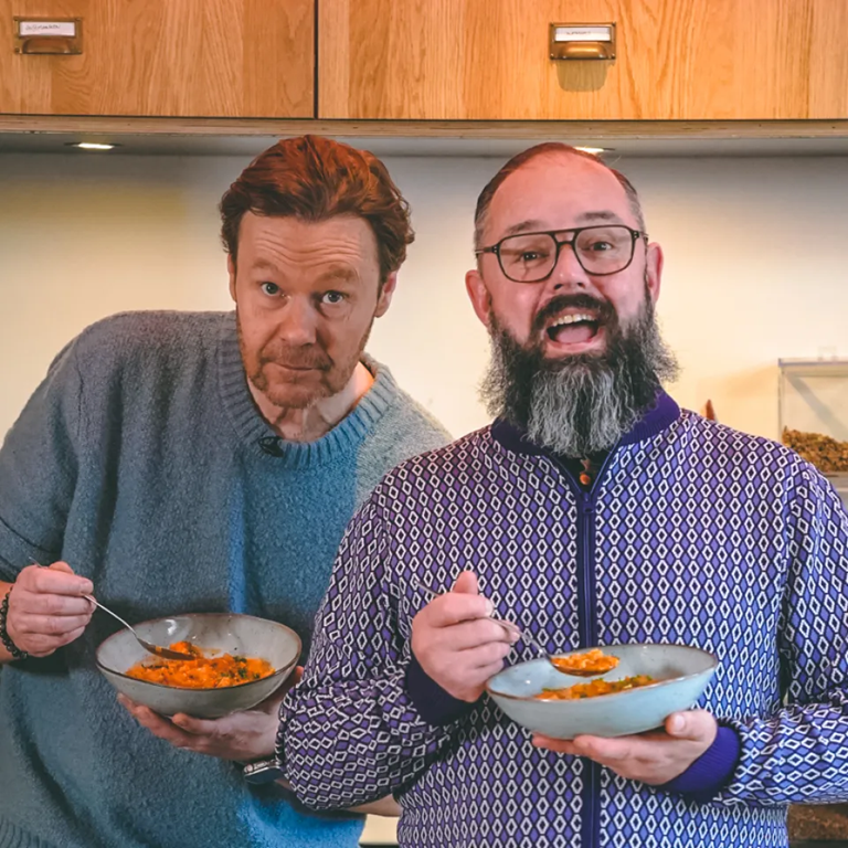 Twee mannen in een keuken, met kommen en lepels in hun handen, glimlachend. De ene draagt een blauwe trui, de andere een vest met rits en een patroon. Eten in kommen.