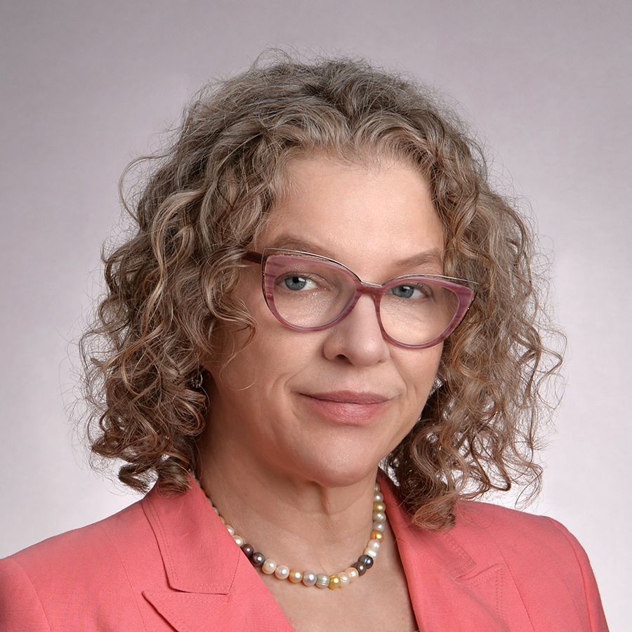 A person with curly hair and glasses is wearing a pink jacket and pearl necklace.