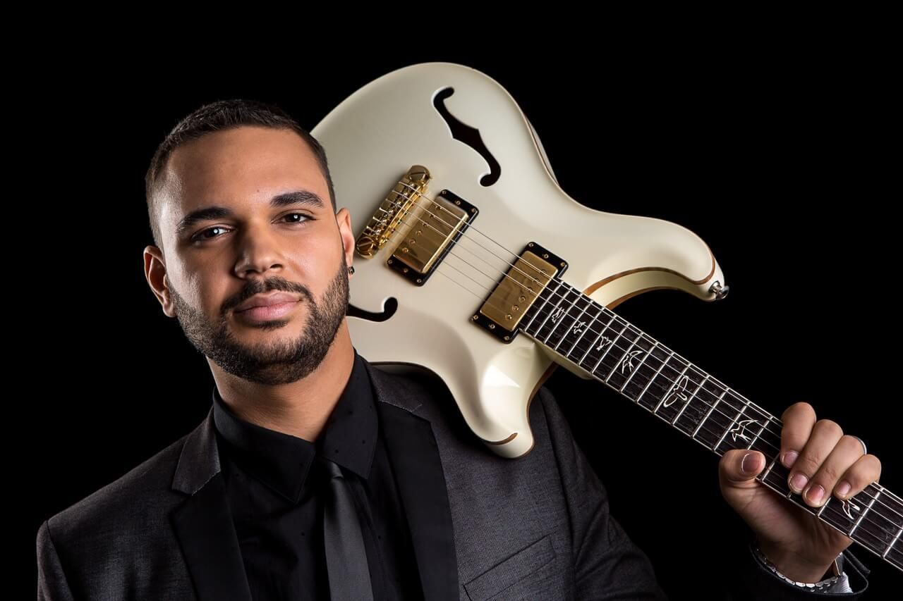 Man in black suit holding a white electric guitar against a black background