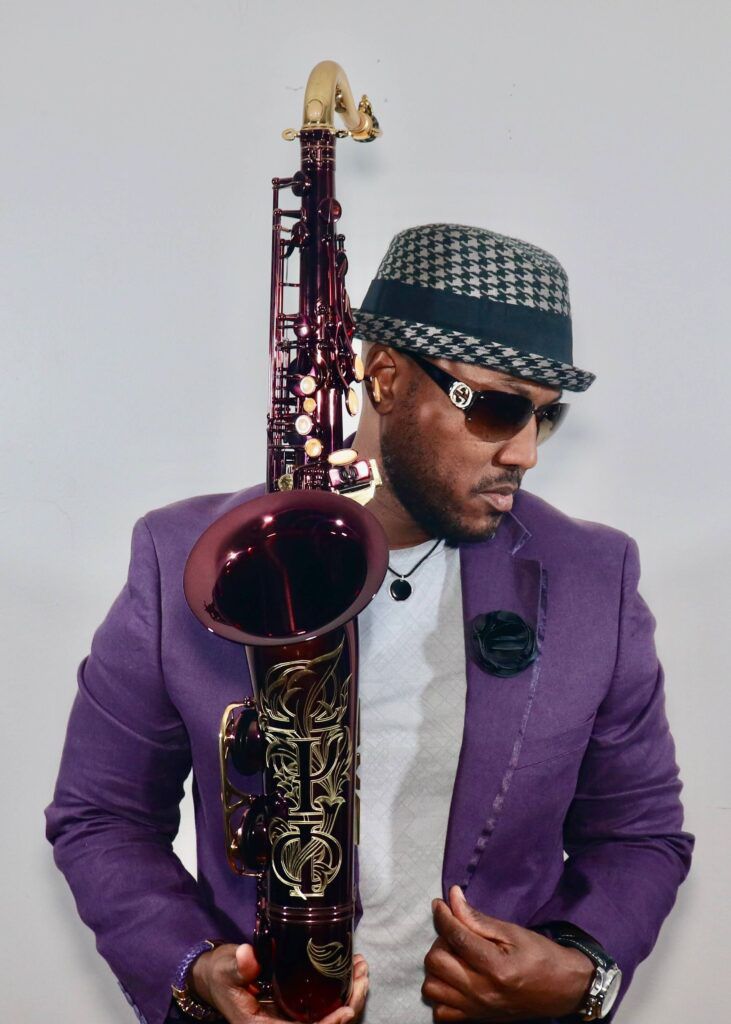 Man in purple suit and patterned hat holding a saxophone, posed against a white background.