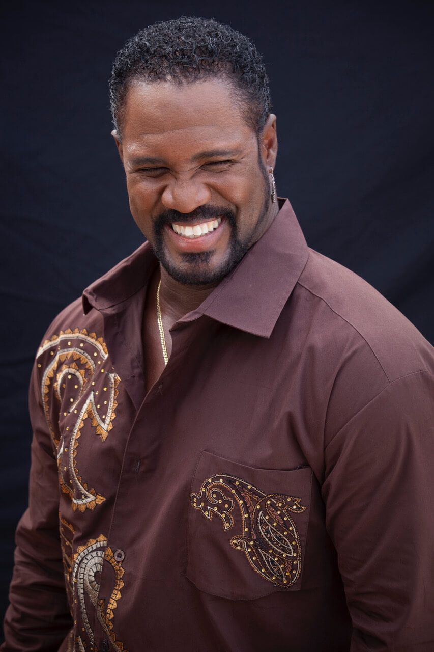 Smiling man in a brown shirt with gold embroidery against a dark background