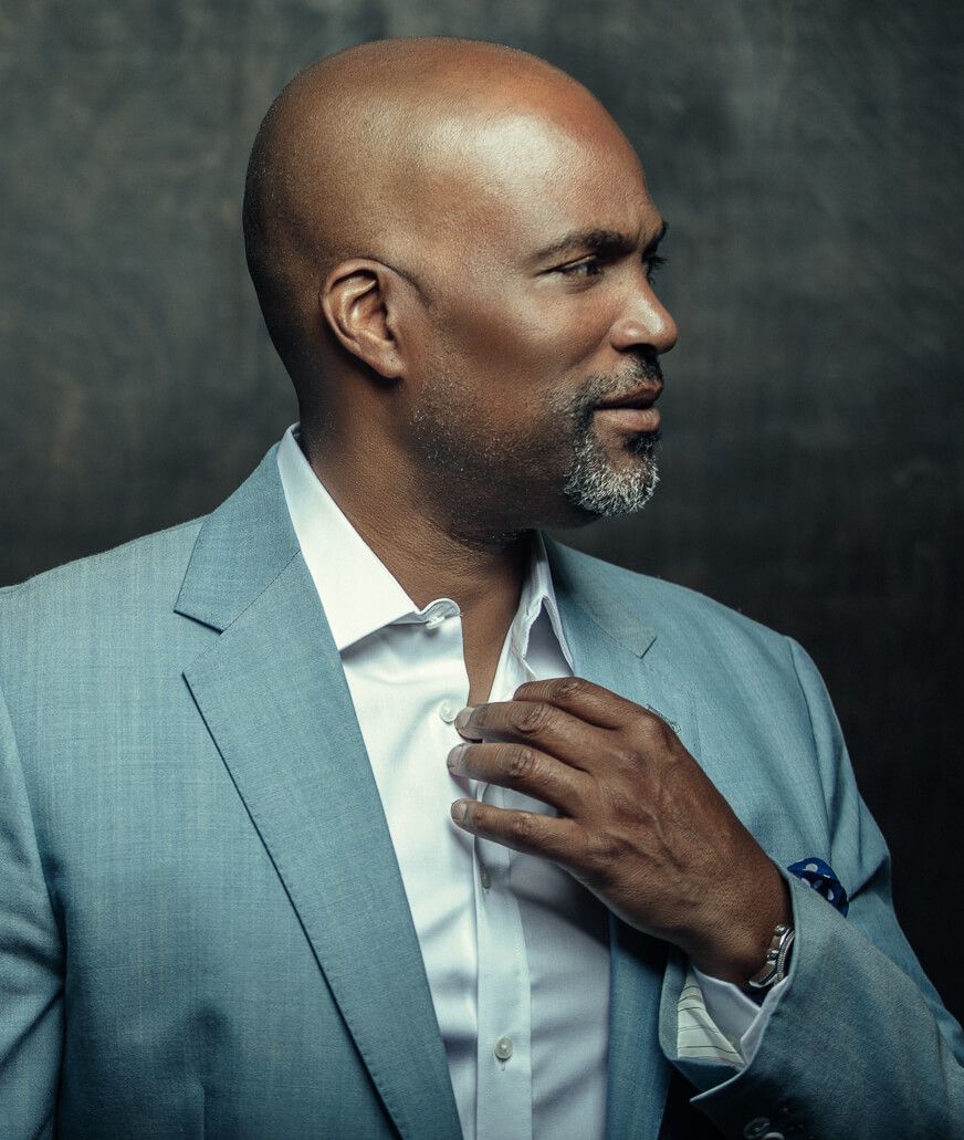 Portrait of a bald man in a light blue suit adjusting his collar against a dark studio background