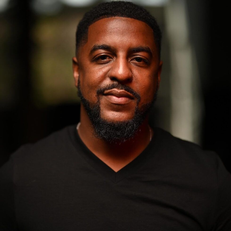 Portrait of a man in a black shirt against a blurred dark background, smiling softly