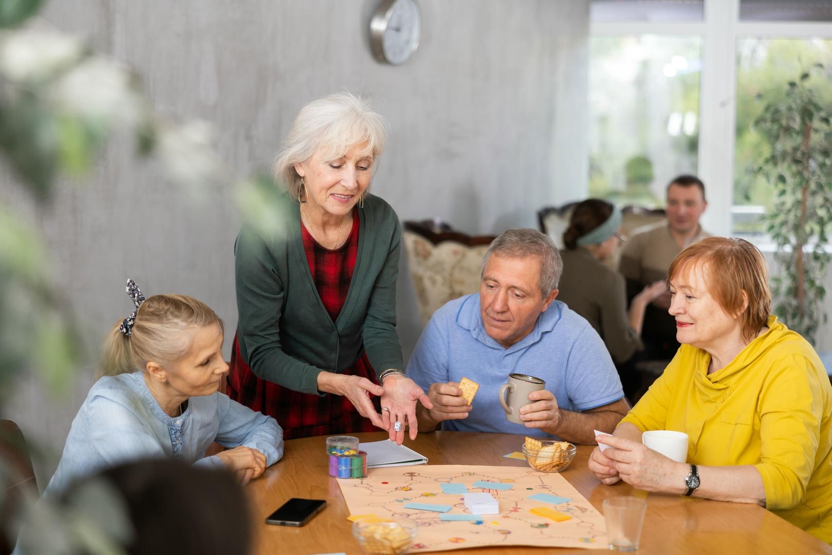 Group of people playing a board game at a table. Woman explains rules. Others watch, eat snacks.