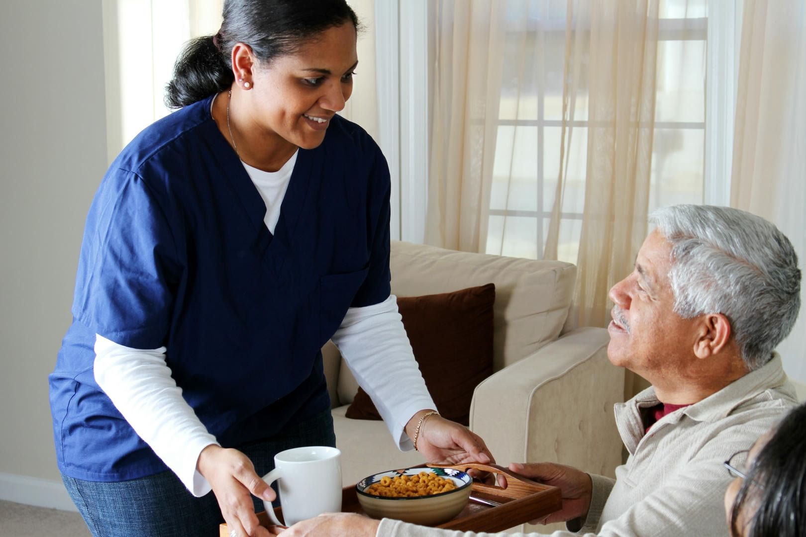 Caregiver serving a meal to an elderly man at home; both smiling.