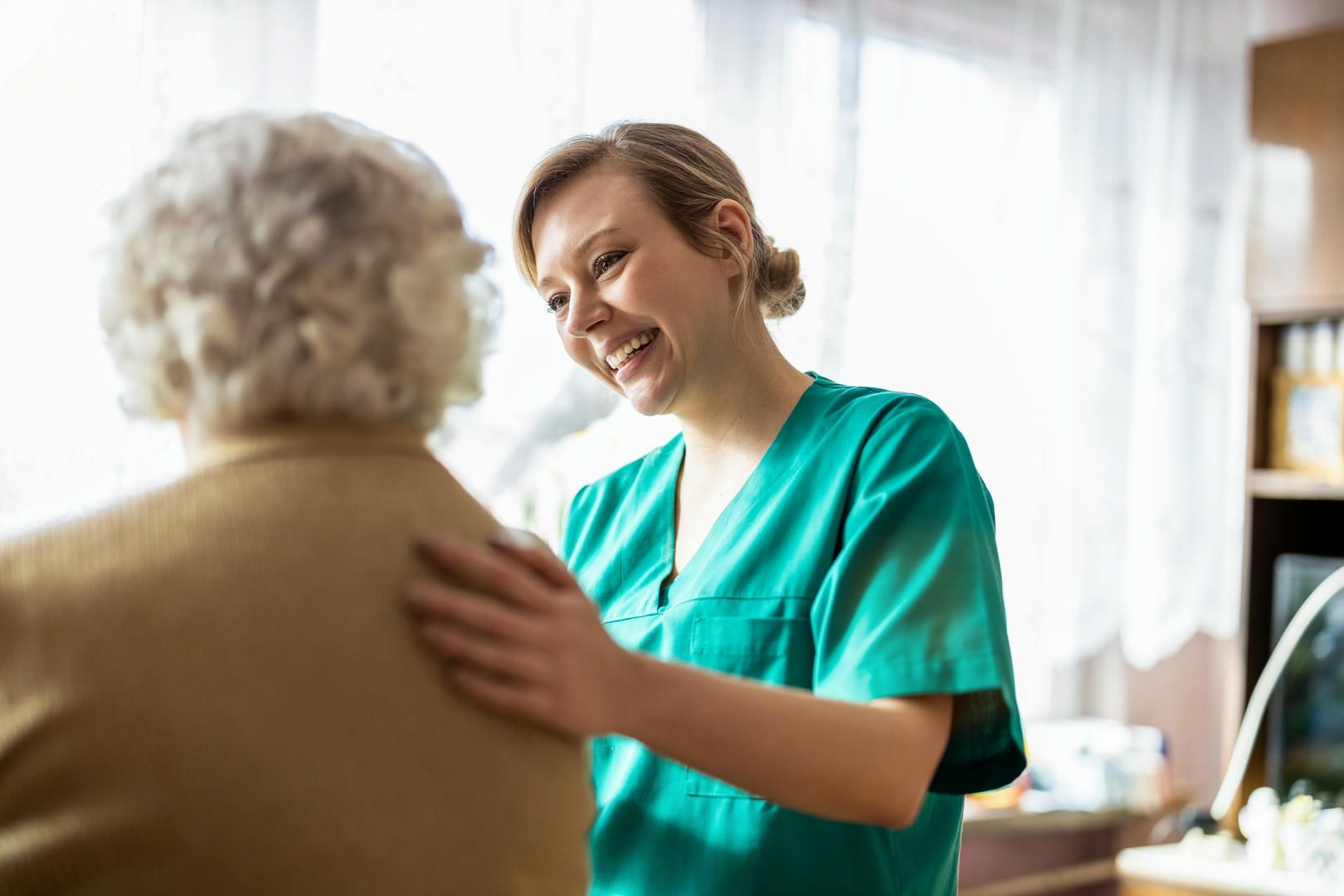Smiling nurse in green scrubs comforts an elderly woman.