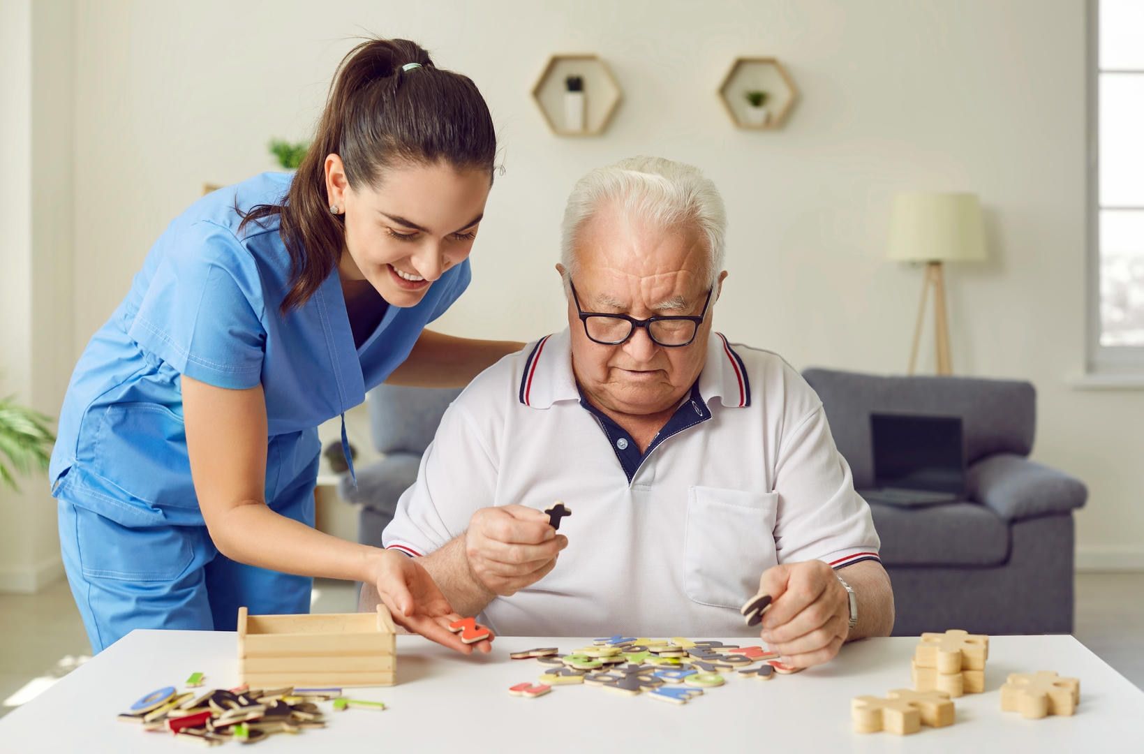 Caregiver helping an elderly man with a jigsaw puzzle at a table; smiling, indoor setting.