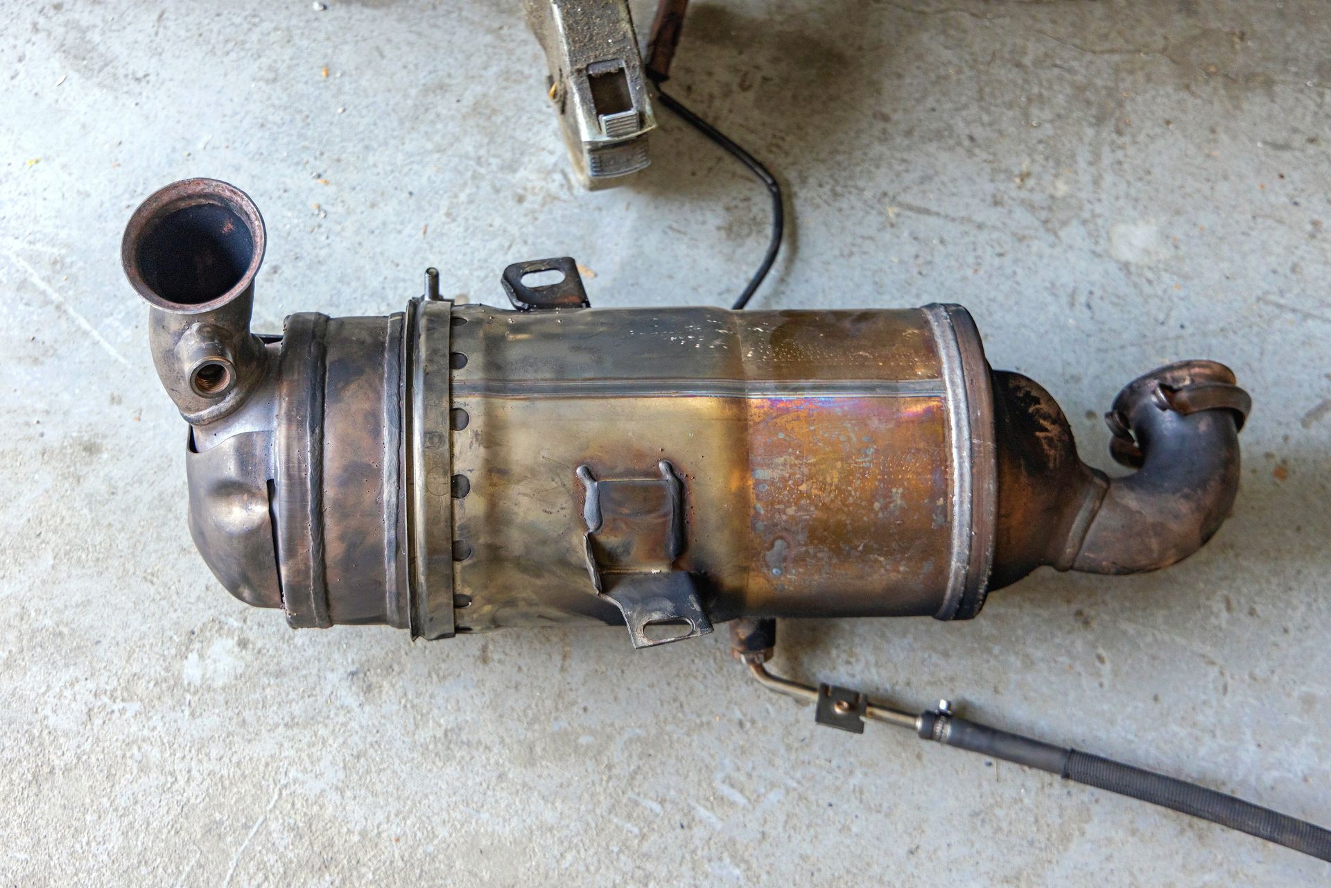 A used, rusty diesel particulate filter with inlet and outlet pipes, likely for a vehicle, on a concrete surface.