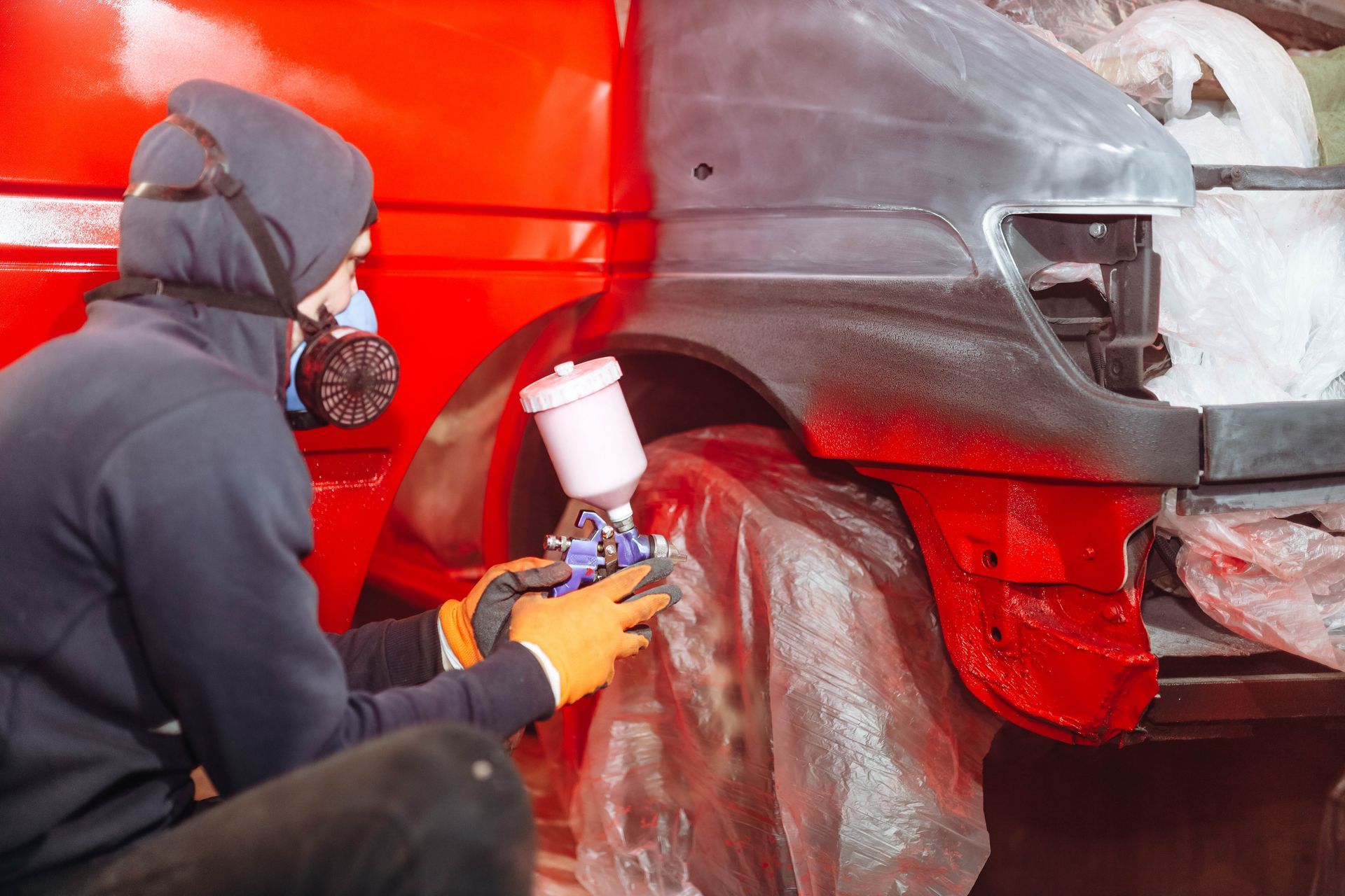 Person in protective gear spray-painting a red vehicle fender with a spray gun.