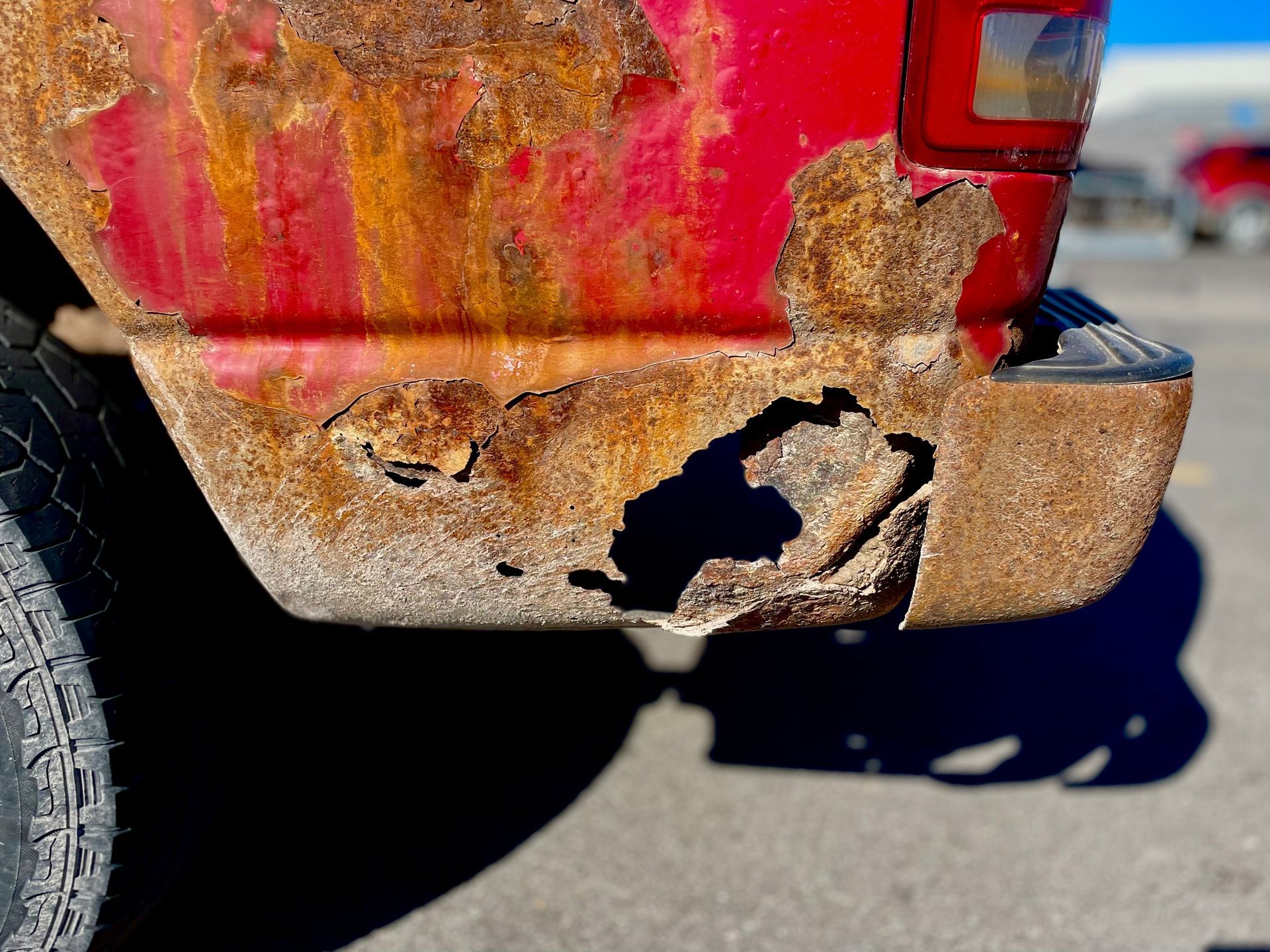 Heavily rusted red truck bed, with large holes and damaged bumper in a sunny outdoor setting.