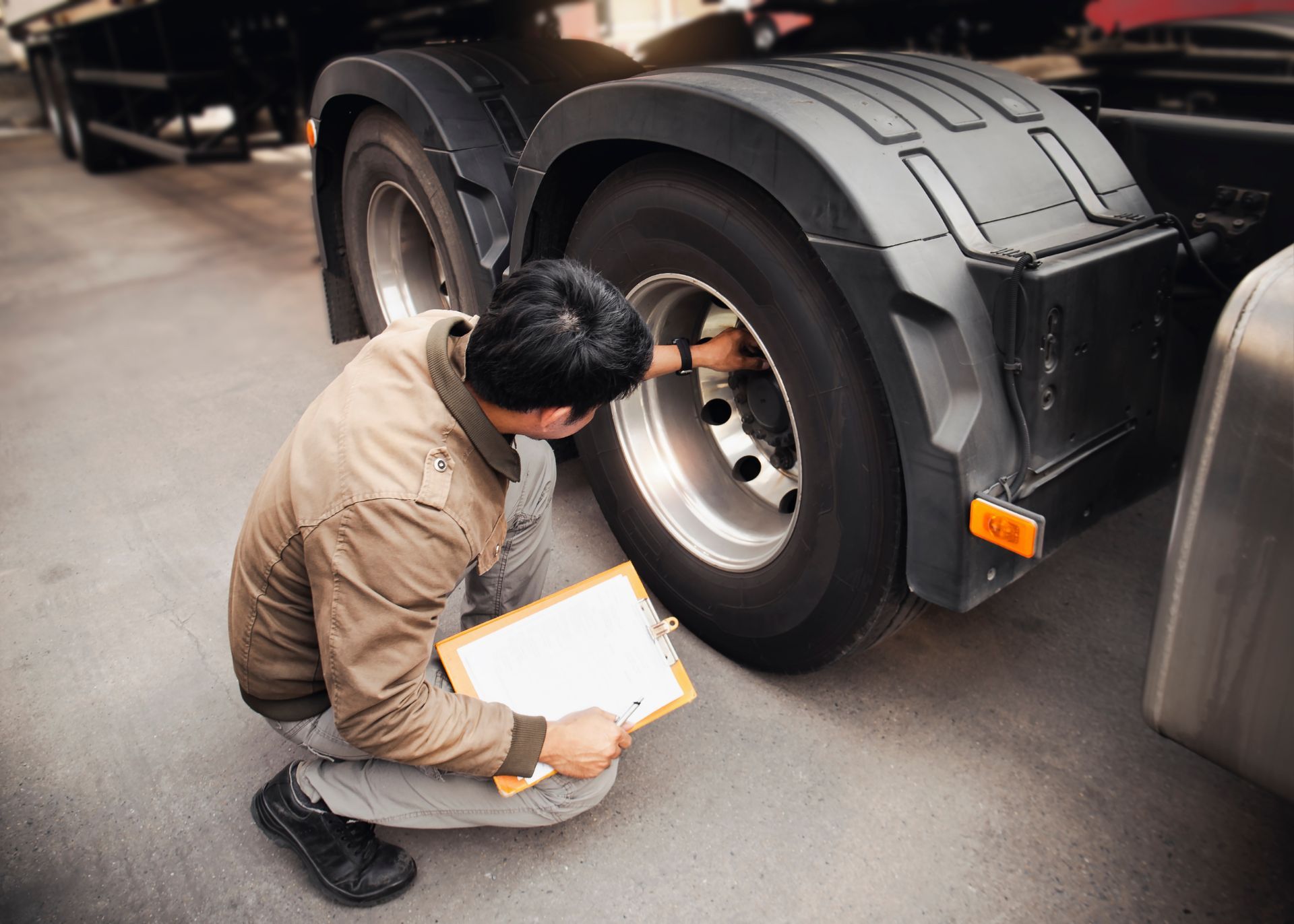Person inspecting truck tire, holding a clipboard in a garage.
