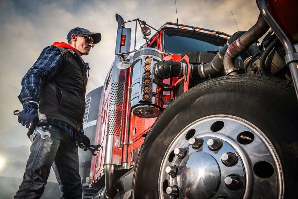 Trucker checking his red semi-truck's tire. He wears a vest, hat, and holds a wrench outside.