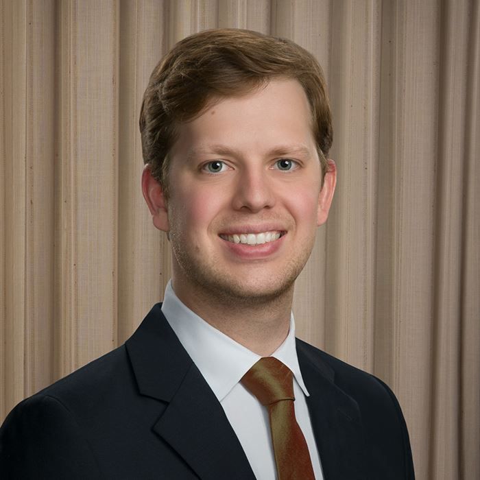 Smiling man in a suit and tie, facing the camera, against a wood-paneled background.