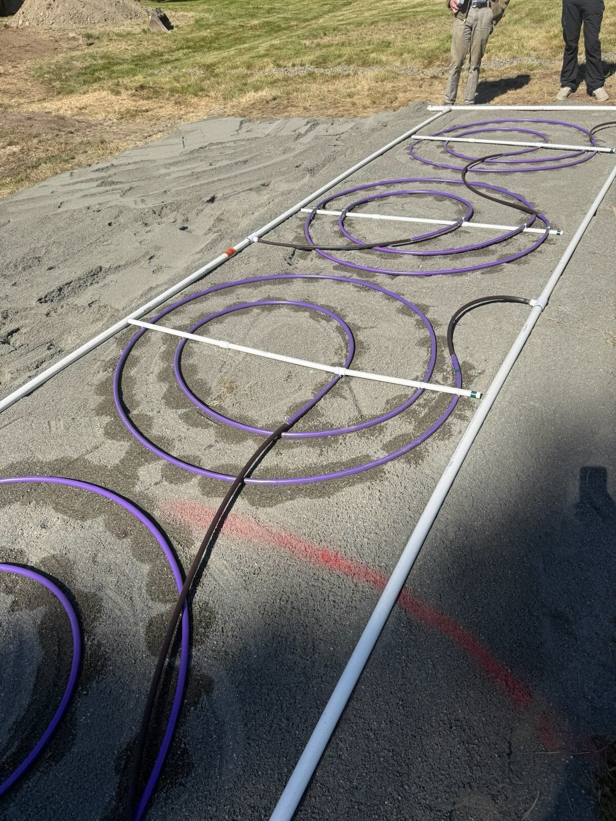 Purple tubing is coiled into circles and connected by white PVC pipes on a gravel ground with two people standing nearby.