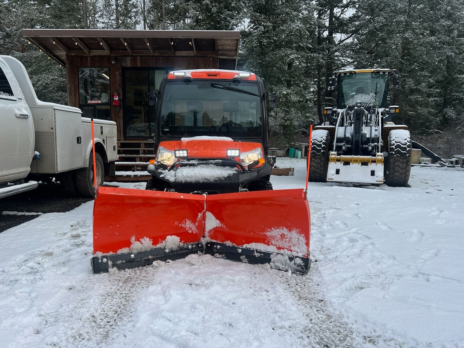 An orange UTV with a snowplow parked in a snow-covered area, with a white truck and heavy machinery nearby.