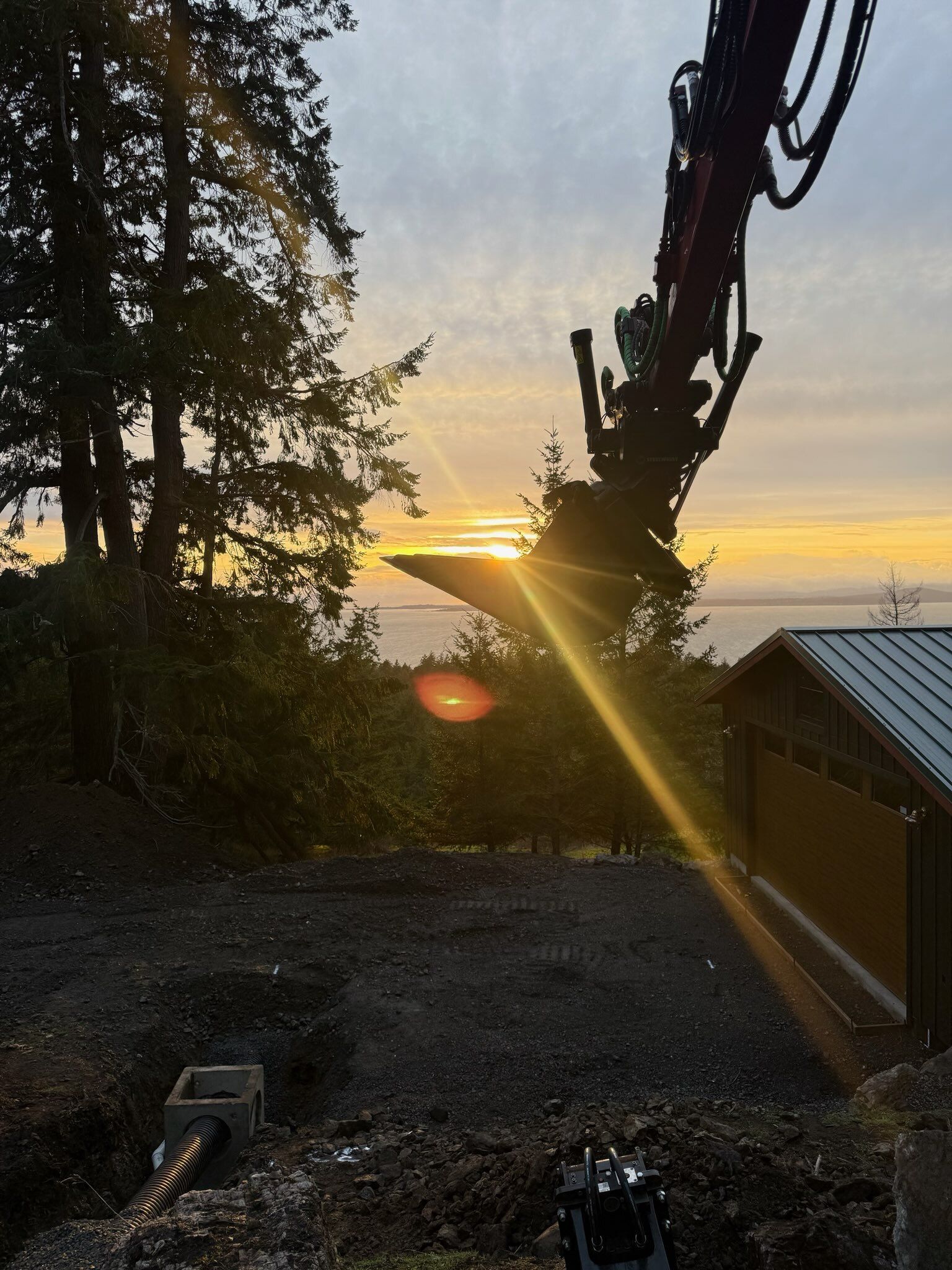 A construction crane arm silhouettes against a vibrant sunset over a treelined landscape and a nearby building.