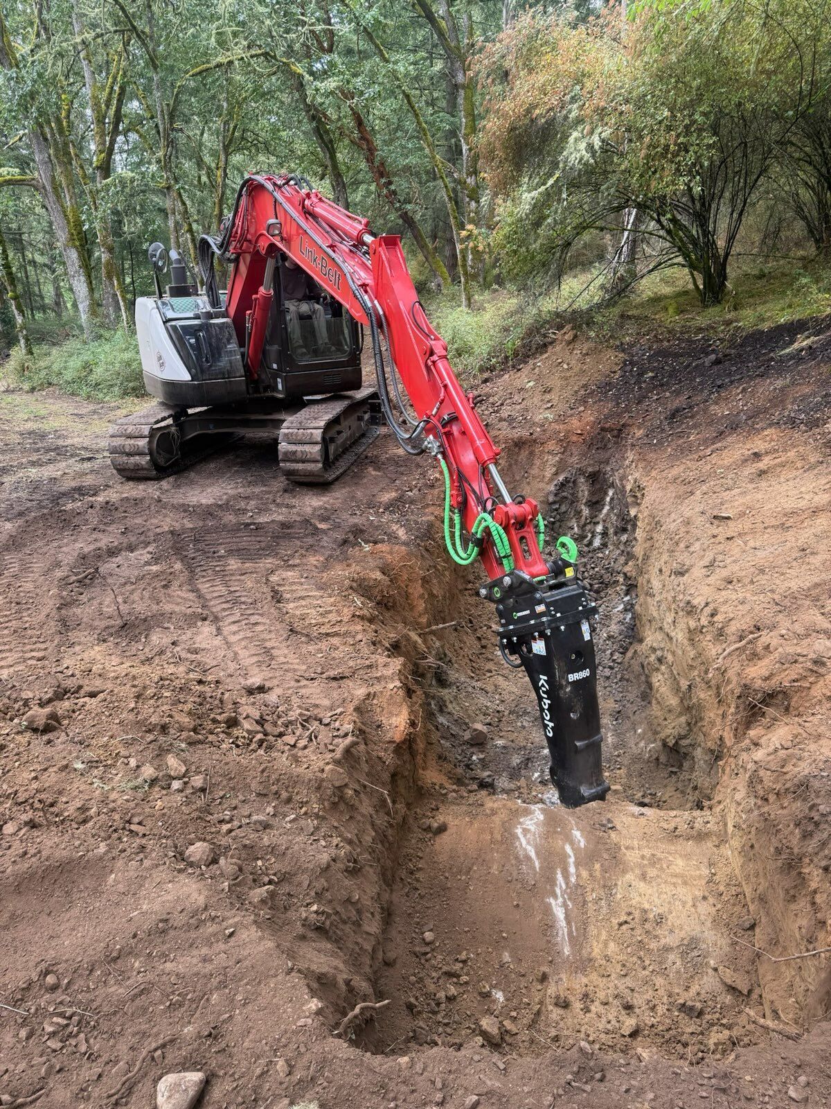 A red and white excavator with a hydraulic breaker attachment works in a dirt trench within a wooded area.