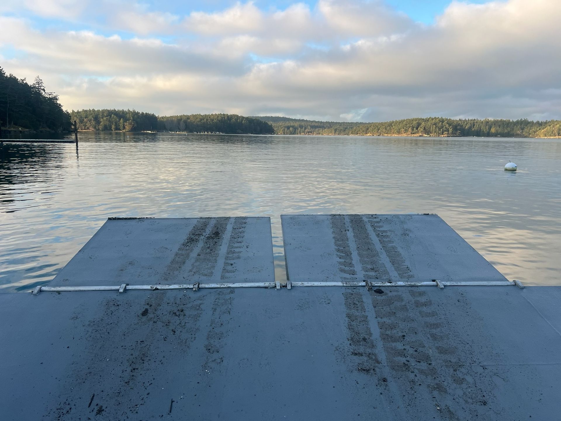 A view from a grey metal ferry ramp extending over calm water toward a forested shoreline under a cloudy sky.
