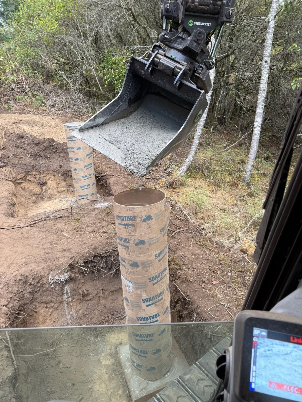 An excavator bucket pours wet concrete into a Sonotube cylindrical form set in the ground at a construction site.