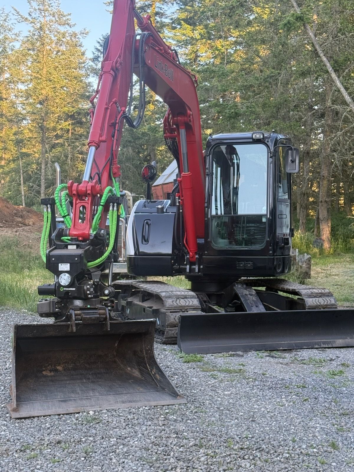 A red and black tracked excavator with a large metal bucket parked on a gravel surface near a treeline.